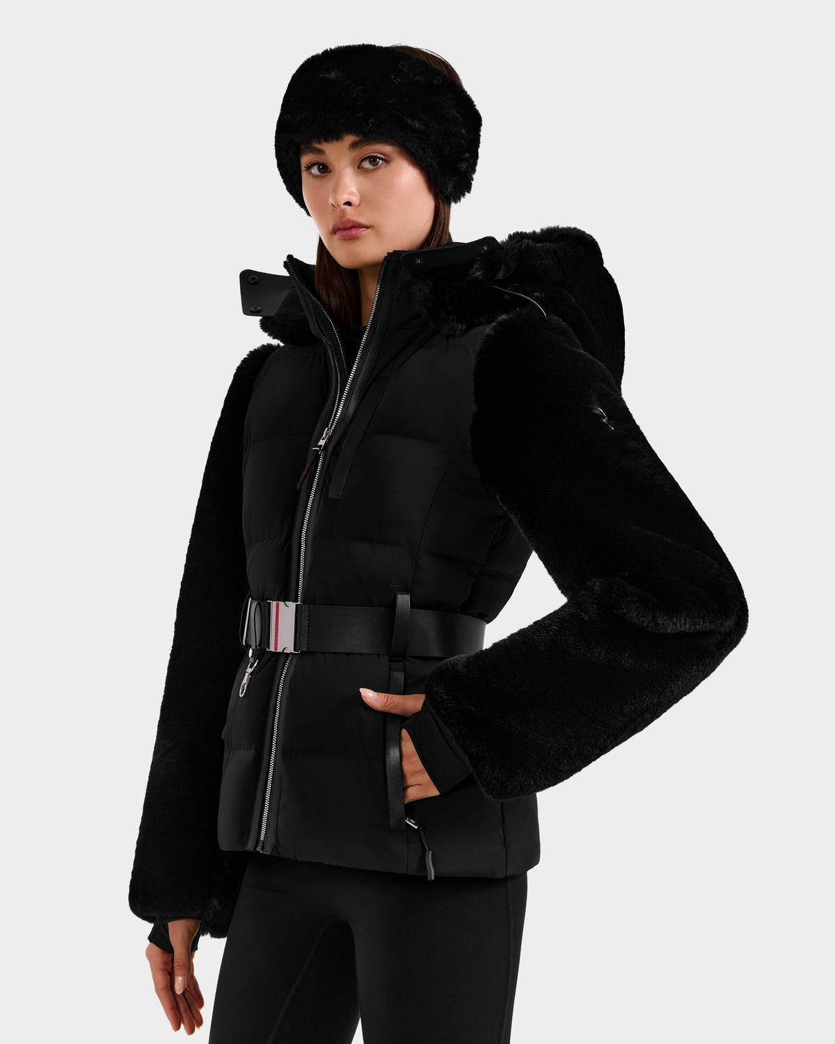 A woman stands against a plain light gray background wearing the Rudsak JOANNA F - 8125593 Black belted faux-fur ski jacket with hood and a matching black faux fur headband.