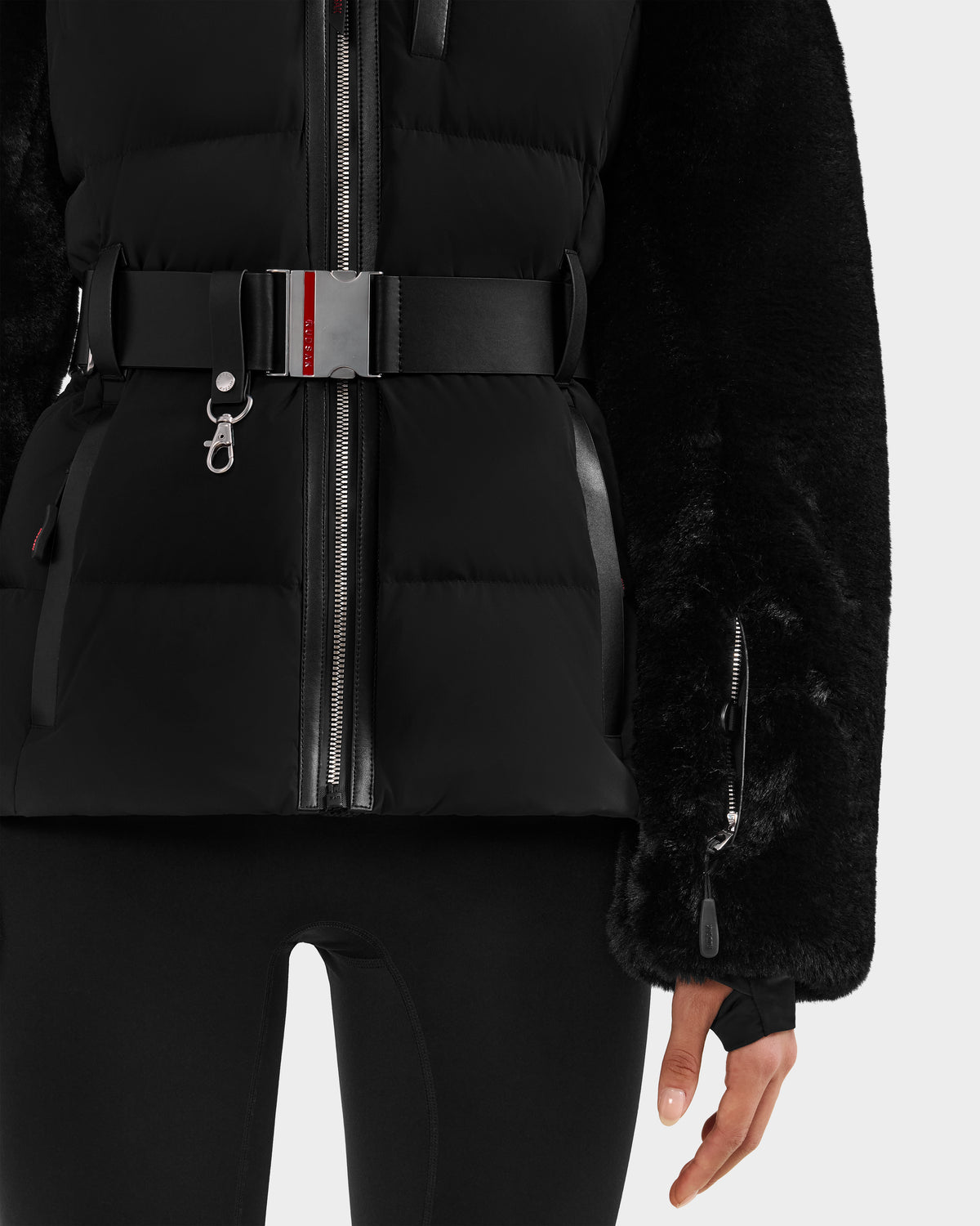 A person is wearing the Rudsak JOANNA F - 8125593 Black women's down ski jacket with a silver zipper, matching belt, and textured sleeves, paired with black leggings. Only the torso and upper legs are visible.