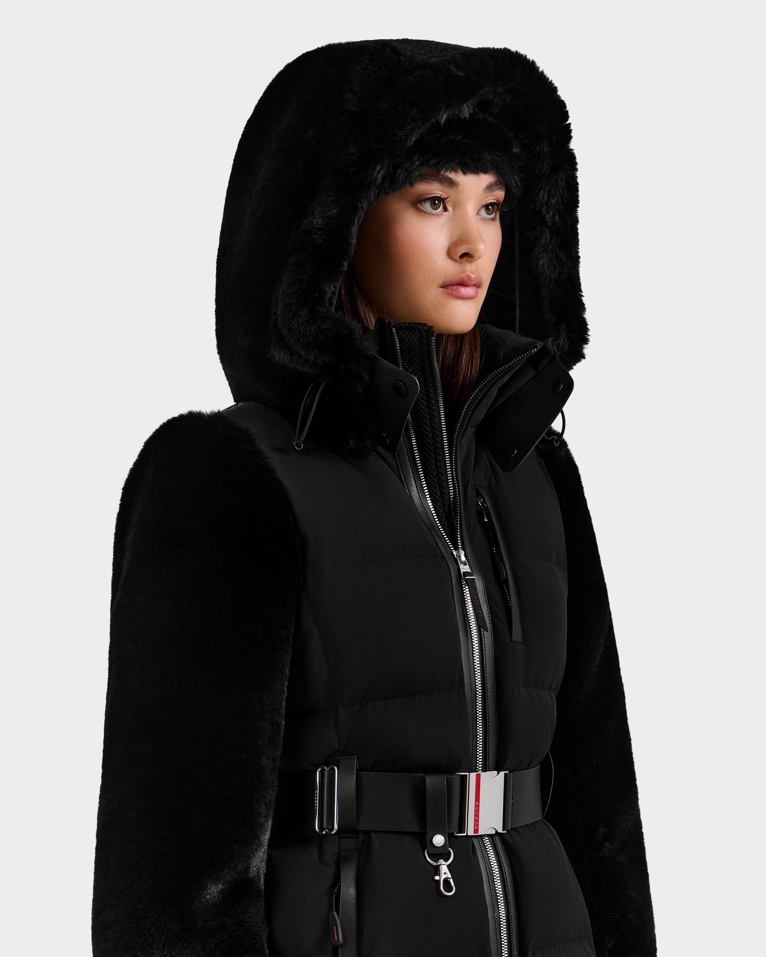 A woman models the Rudsak JOANNA F - 8125593 Black ski jacket featuring faux fur, a hood, high collar, and belted waist—combining style and warmth for winter adventures against a plain light background.
