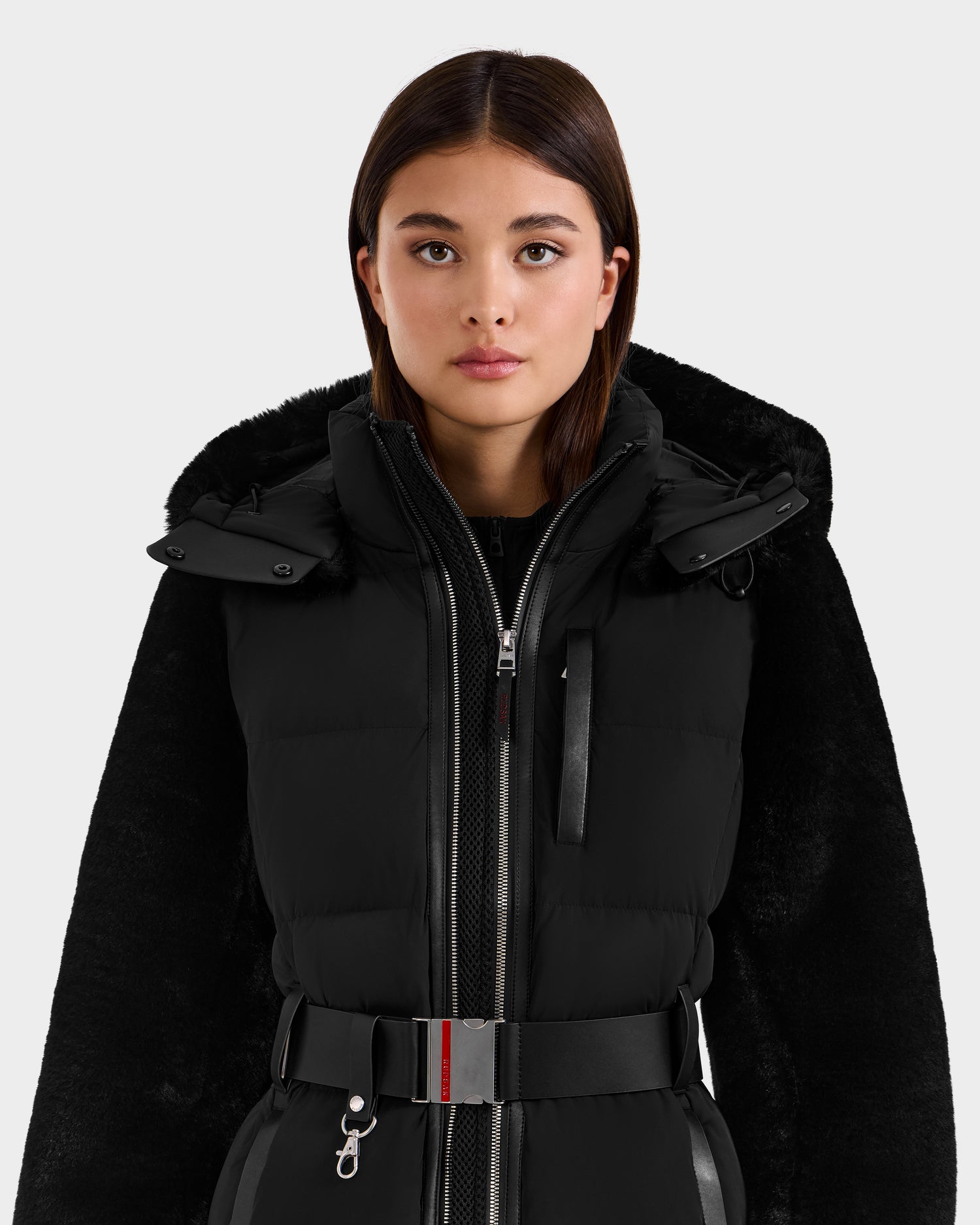 A young woman with straight brown hair models the Rudsak JOANNA F - 8125593 Black women’s down ski jacket with a belted waist, standing against a plain light background and looking directly at the camera.