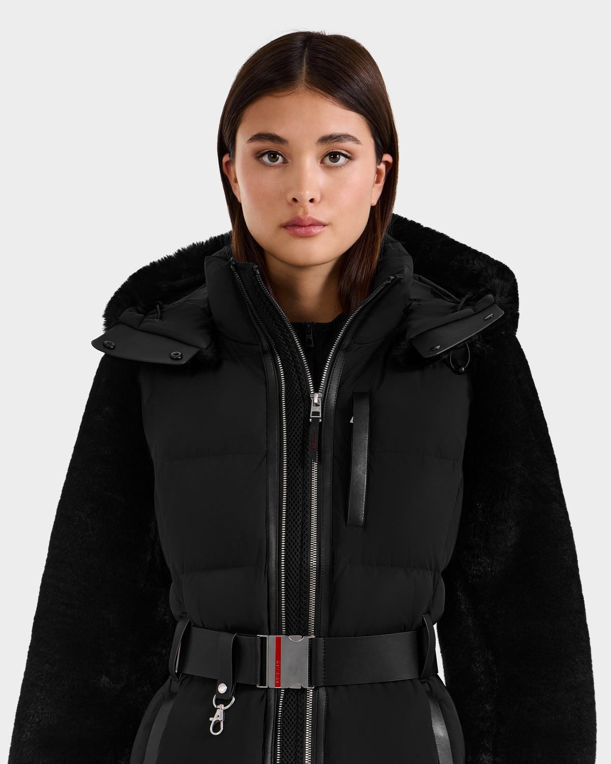 A young woman with straight brown hair models the Rudsak JOANNA F - 8125593 Black women’s down ski jacket with a belted waist, standing against a plain light background and looking directly at the camera.