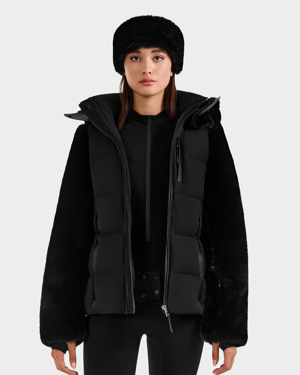 A woman stands facing forward against a plain light gray background wearing the Rudsak JOANNA F - 8125593 Black faux-fur ski jacket with RECCO technology, a furry hat, and black pants.