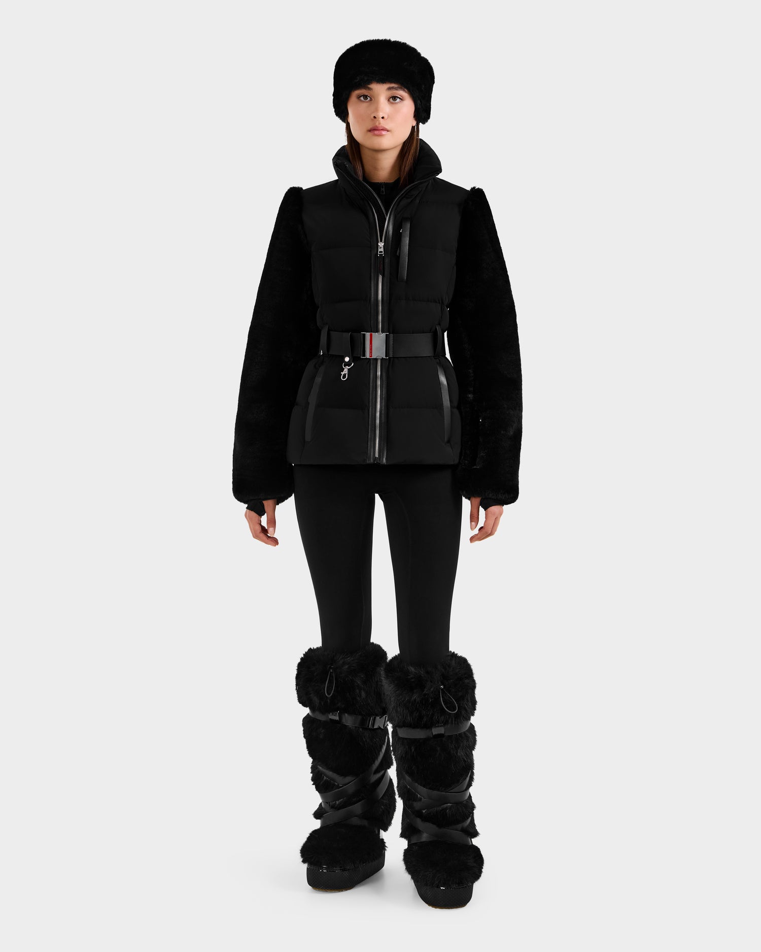 A woman faces forward against a white background, wearing the Rudsak JOANNA F - 8125593 Black women’s down ski jacket with fur sleeves, fitted pants, and large fur-trimmed boots.