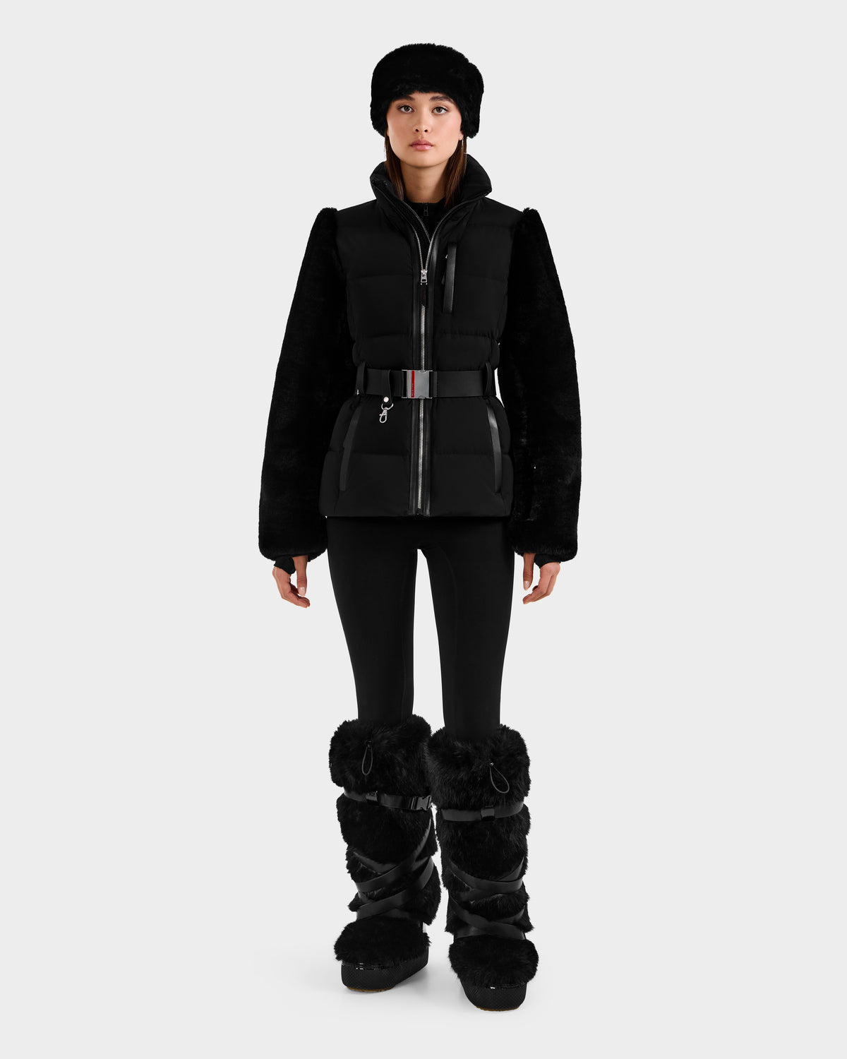 A woman faces forward against a white background, wearing the Rudsak JOANNA F - 8125593 Black women’s down ski jacket with fur sleeves, fitted pants, and large fur-trimmed boots.