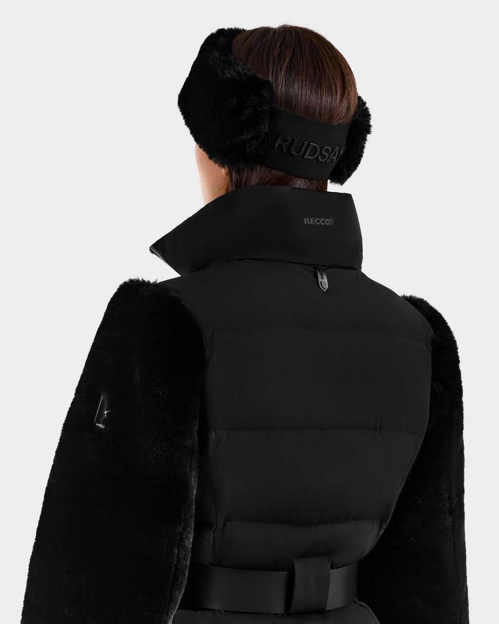 A person is shown from the back wearing the Rudsak JOANNA F - 8125593 Black women’s down ski jacket with a high collar, black belt, and large branded fur earmuffs. The coat features RECCO rescue technology and stylish winter-ready details.