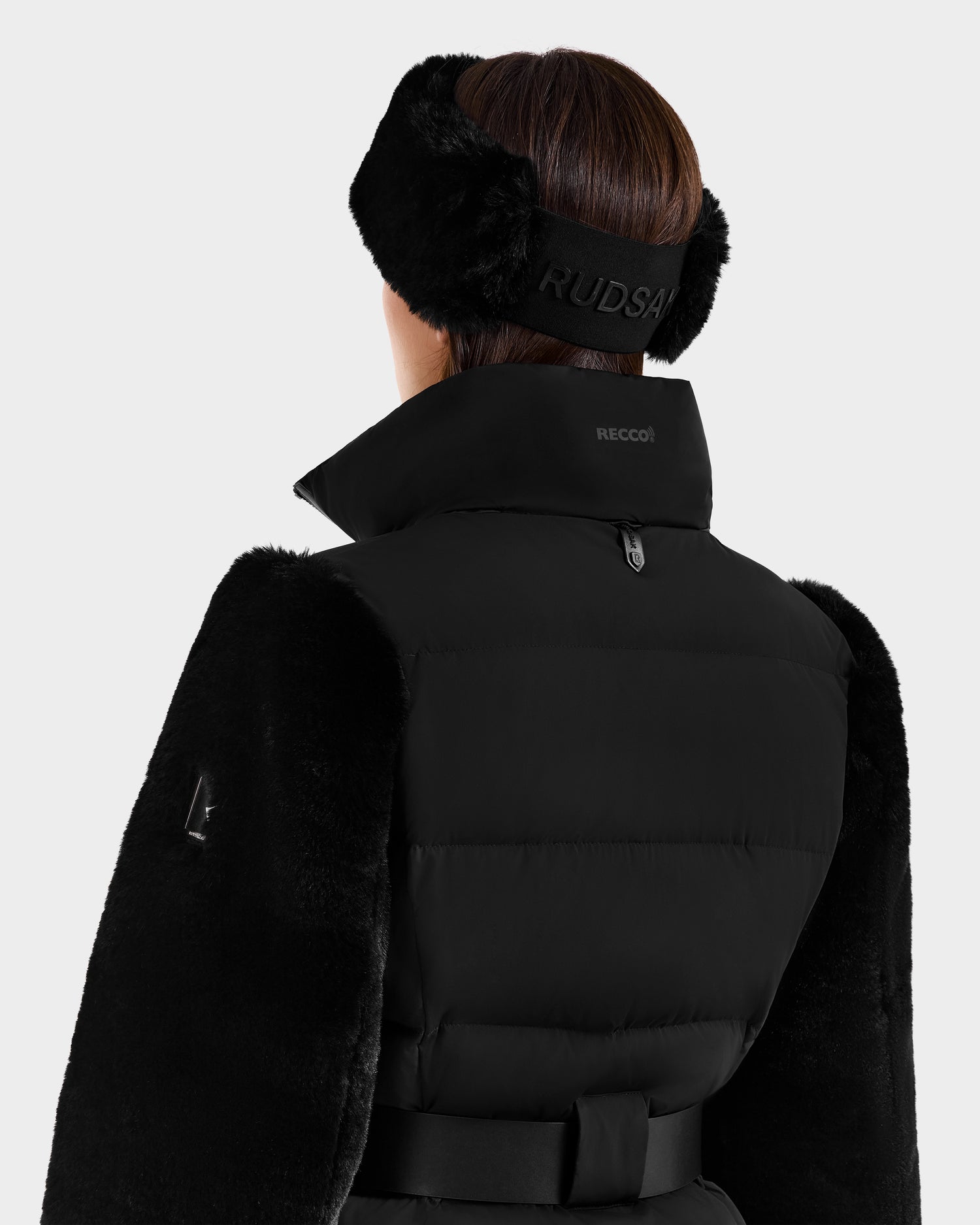 A person is shown from the back wearing the Rudsak JOANNA F - 8125593 Black women’s down ski jacket with a high collar, black belt, and large branded fur earmuffs. The coat features RECCO rescue technology and stylish winter-ready details.