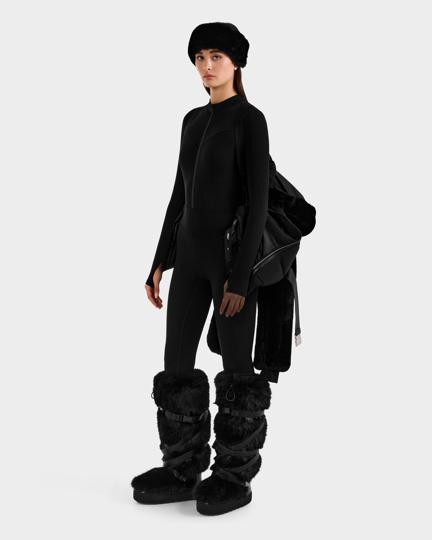 A woman wearing a black fitted bodysuit, black fur hat, and large black furry boots stands against a white background, holding the Rudsak JOANNA F - 8125593 Black women’s down ski jacket with faux-fur trim draped over her arm.