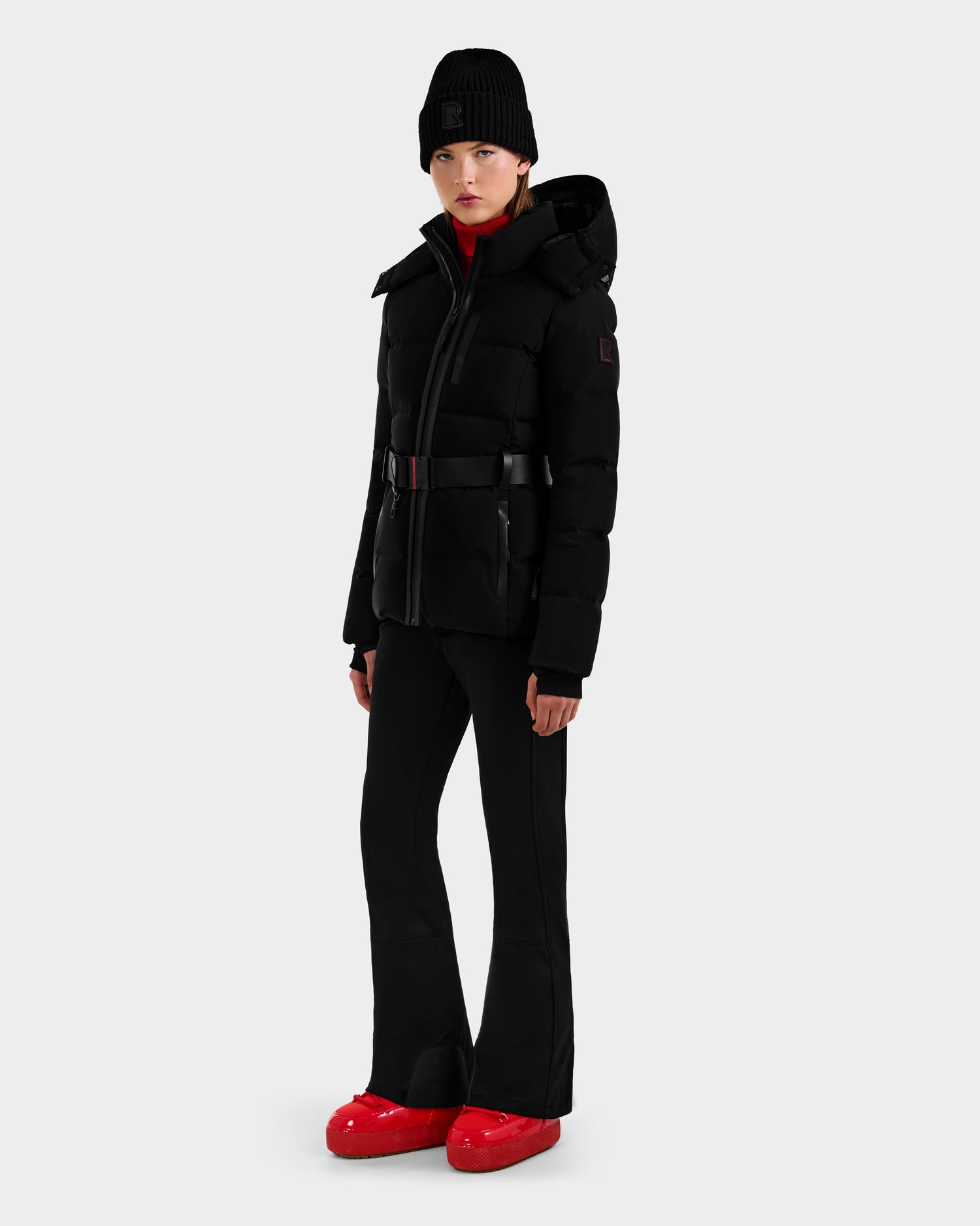 A person stands against a light gray background wearing the Rudsak JOANNA - 8124788 Black women’s down ski jacket with hood, ski-pass pocket, and RECCO rescue tech, paired with black pants, a black beanie, and bright red shoes.