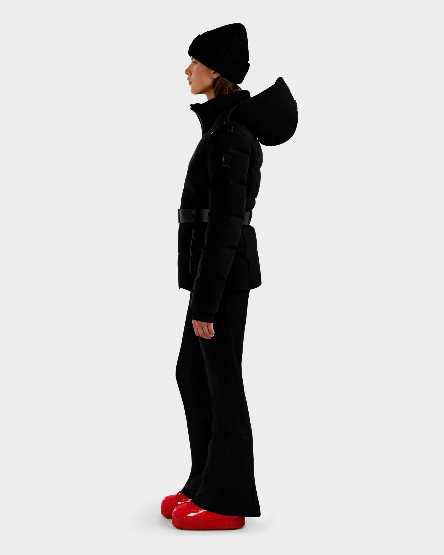 A woman in profile wears Rudsak's JOANNA - 8124788 Black belted women's down ski jacket with RECCO rescue tech, a black beanie, flared black pants, and bright red puffy shoes against a plain background.