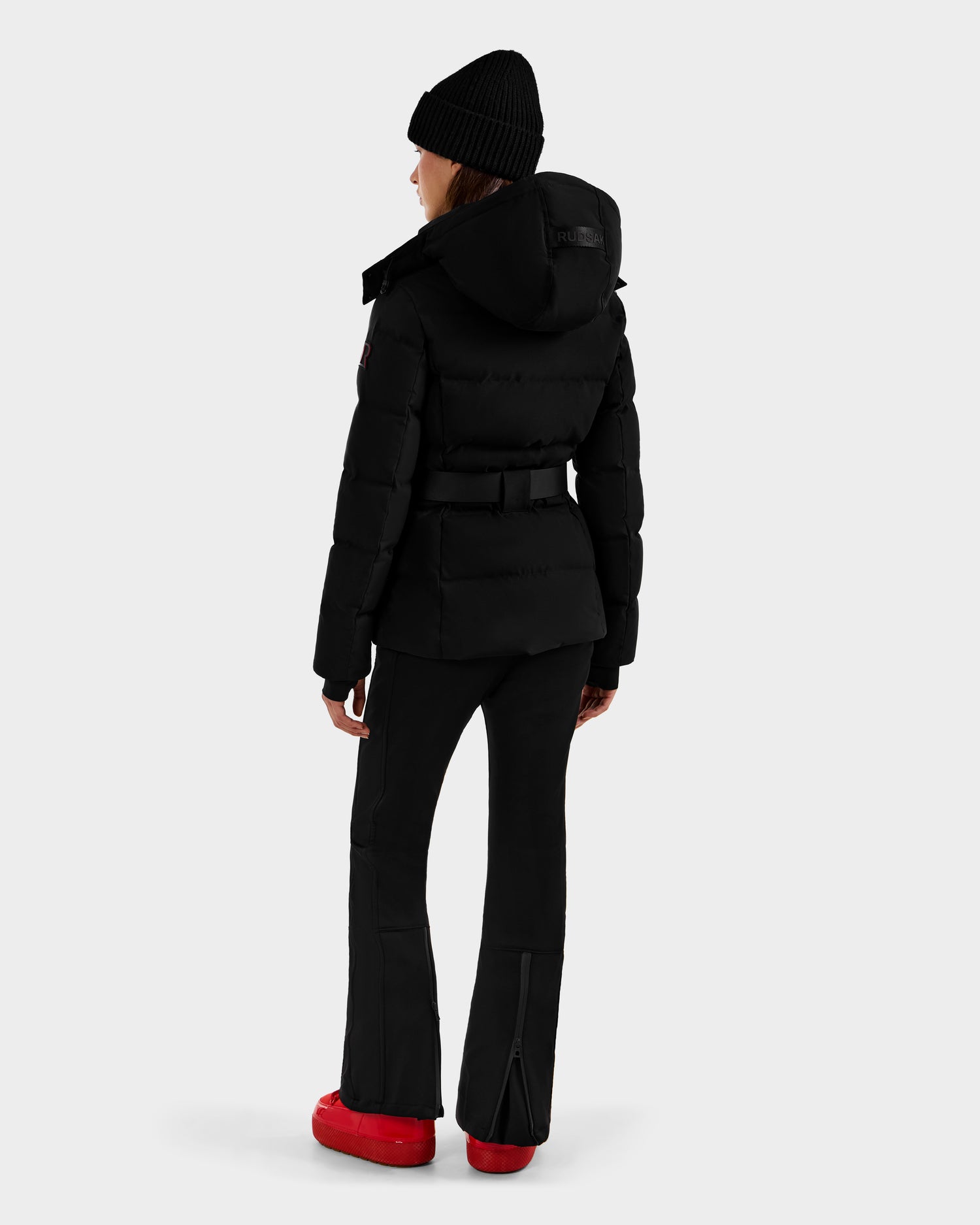 A person wearing the Rudsak JOANNA - 8124788 Black belted women's down ski jacket stands facing away, displaying the back of their winter outfit against a plain light gray background.