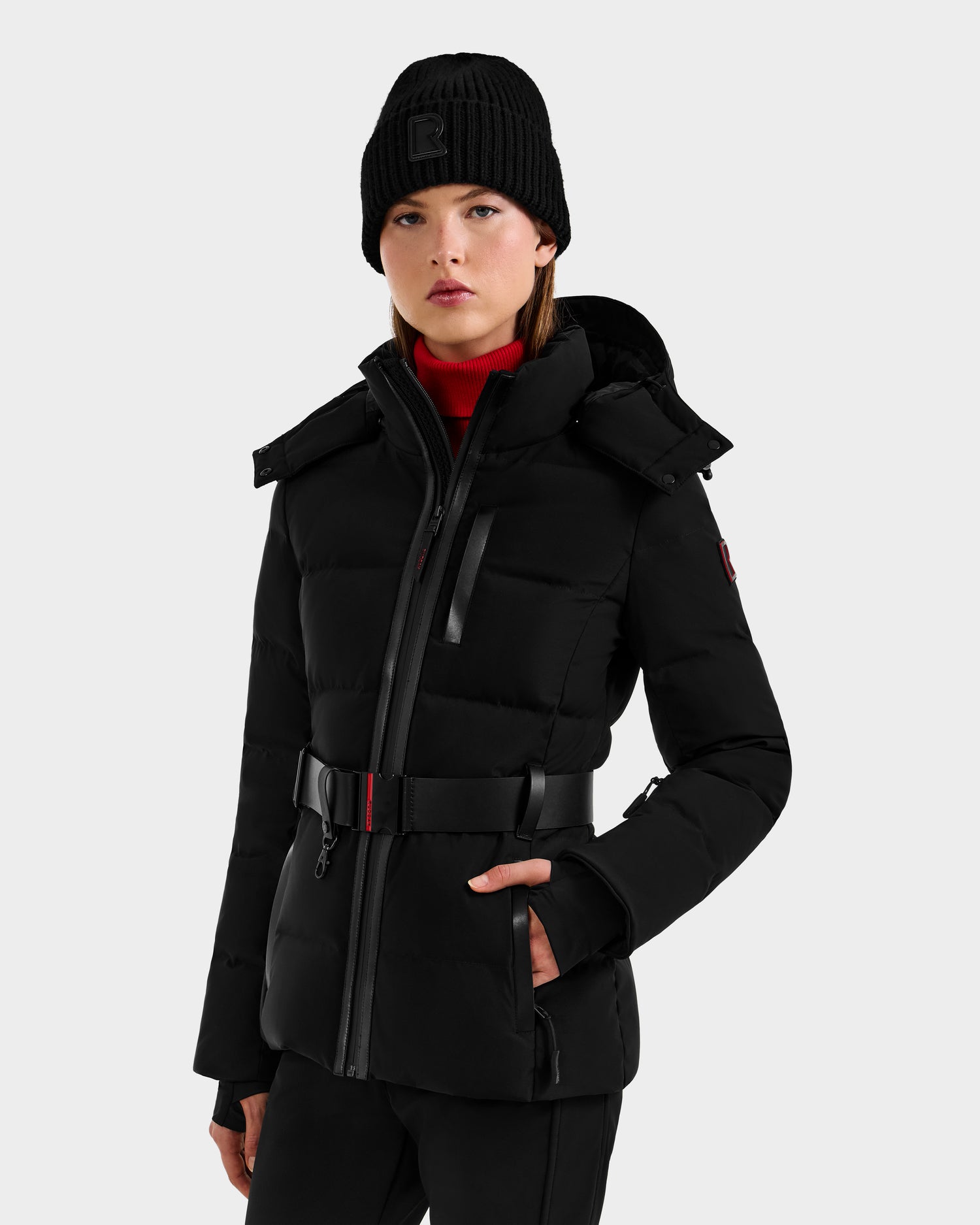 A woman in the Rudsak JOANNA - 8124788 Black belted women’s down ski jacket with a ski-pass pocket, black knit beanie, and red turtleneck stands against a plain light background, looking ahead neutrally.