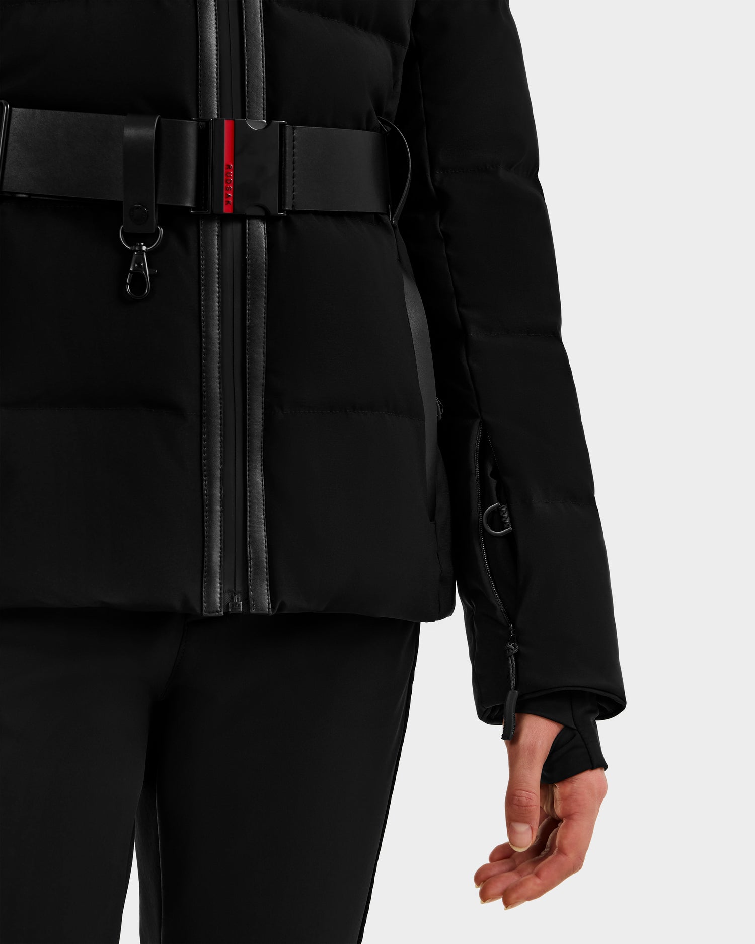 A person wears the Rudsak JOANNA - 8124788 Black belted women's down ski jacket with zip details and matching pants, their right arm relaxed by their side, with only the lower half visible against a light background.