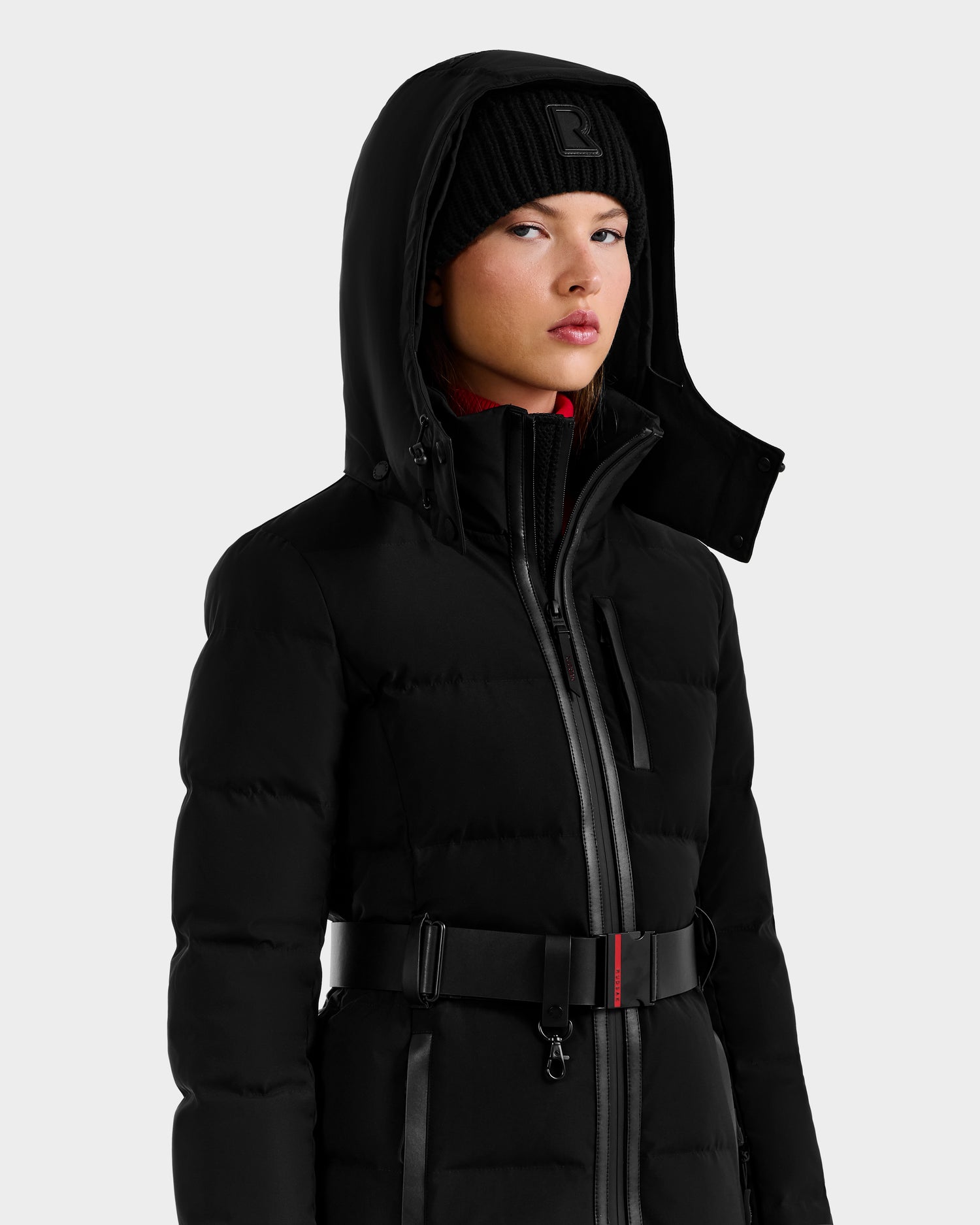 A woman wears the Rudsak JOANNA - 8124788 Black women’s down ski jacket with a belt, a black beanie, and a red inner layer featuring RECCO rescue technology, standing against a light gray background.
