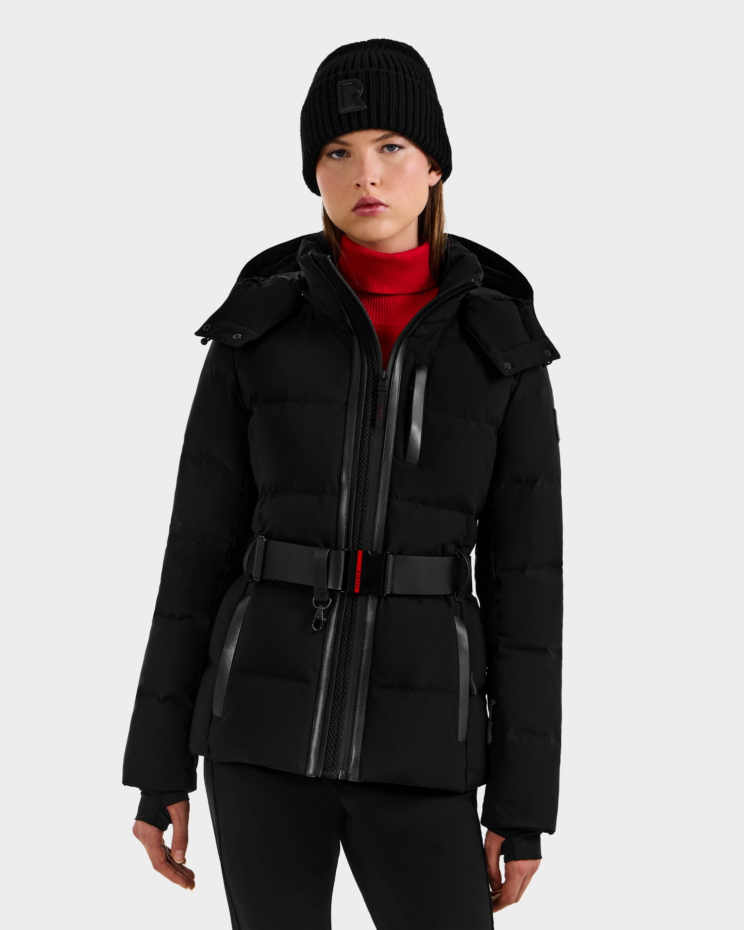 A woman stands facing forward against a light gray background, wearing the Rudsak JOANNA - 8124788 Black women's down ski jacket with a ski-pass pocket, a black knit beanie, and a red turtleneck.