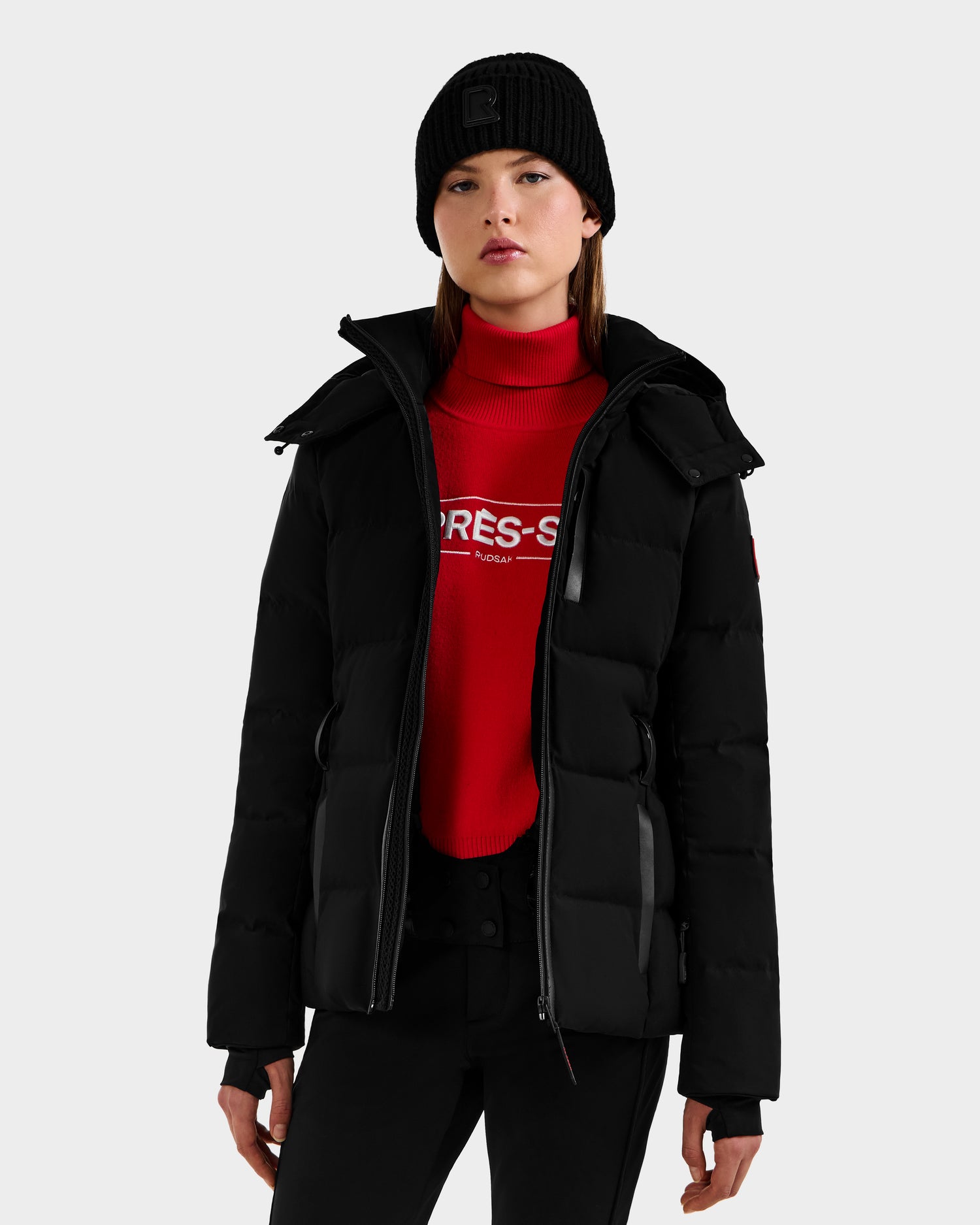 A person wears the Rudsak JOANNA - 8124788 Black women’s down ski jacket with RECCO, a black knit beanie, and a red turtleneck sweater with white text, standing against a light background and looking at the camera neutrally.