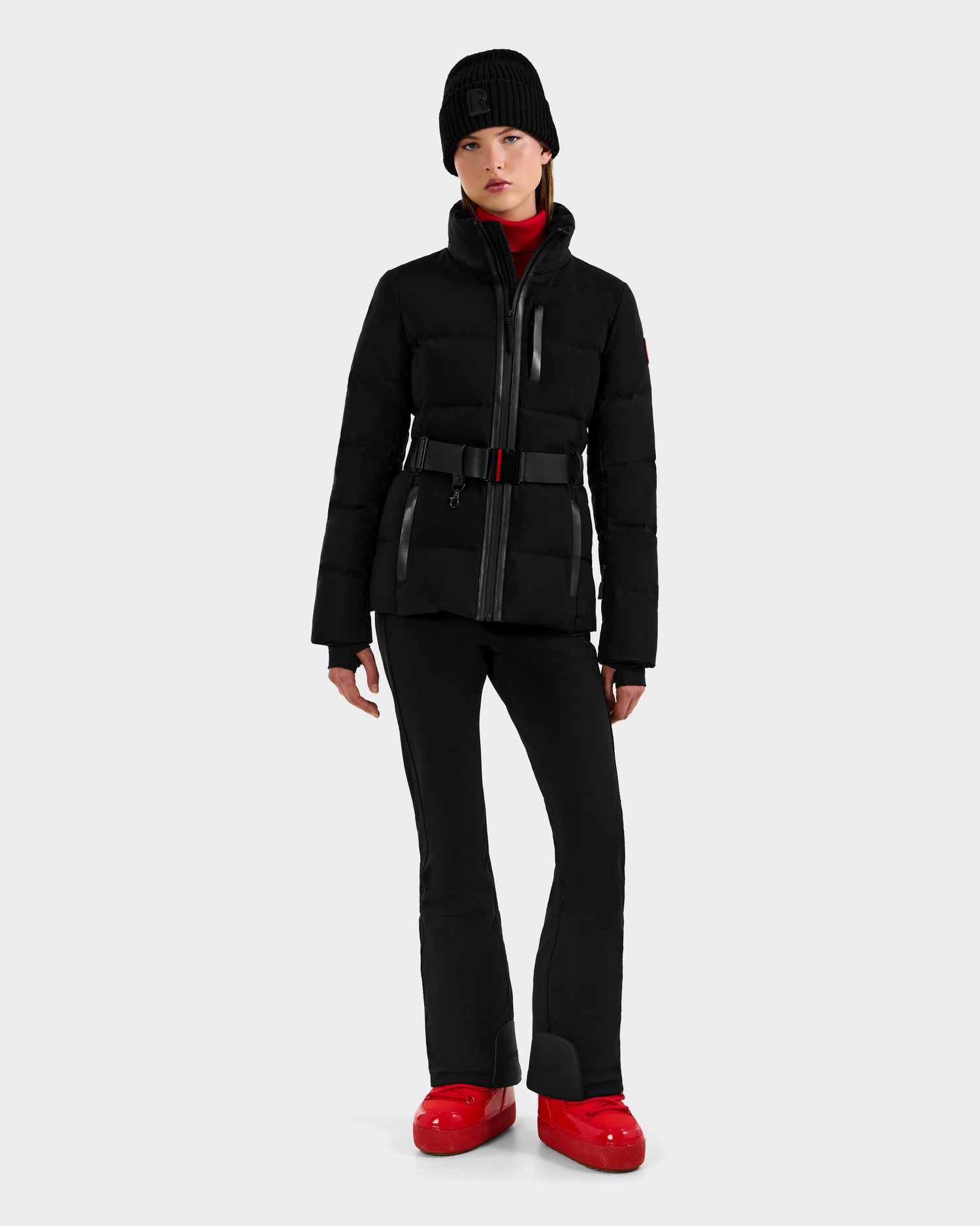 A woman with a neutral expression stands against a light gray background, wearing Rudsak's JOANNA - 8124788 Black women's down ski jacket with a belt and ski-pass pocket, black pants, a black beanie, and red winter boots. Her hands rest by her sides.