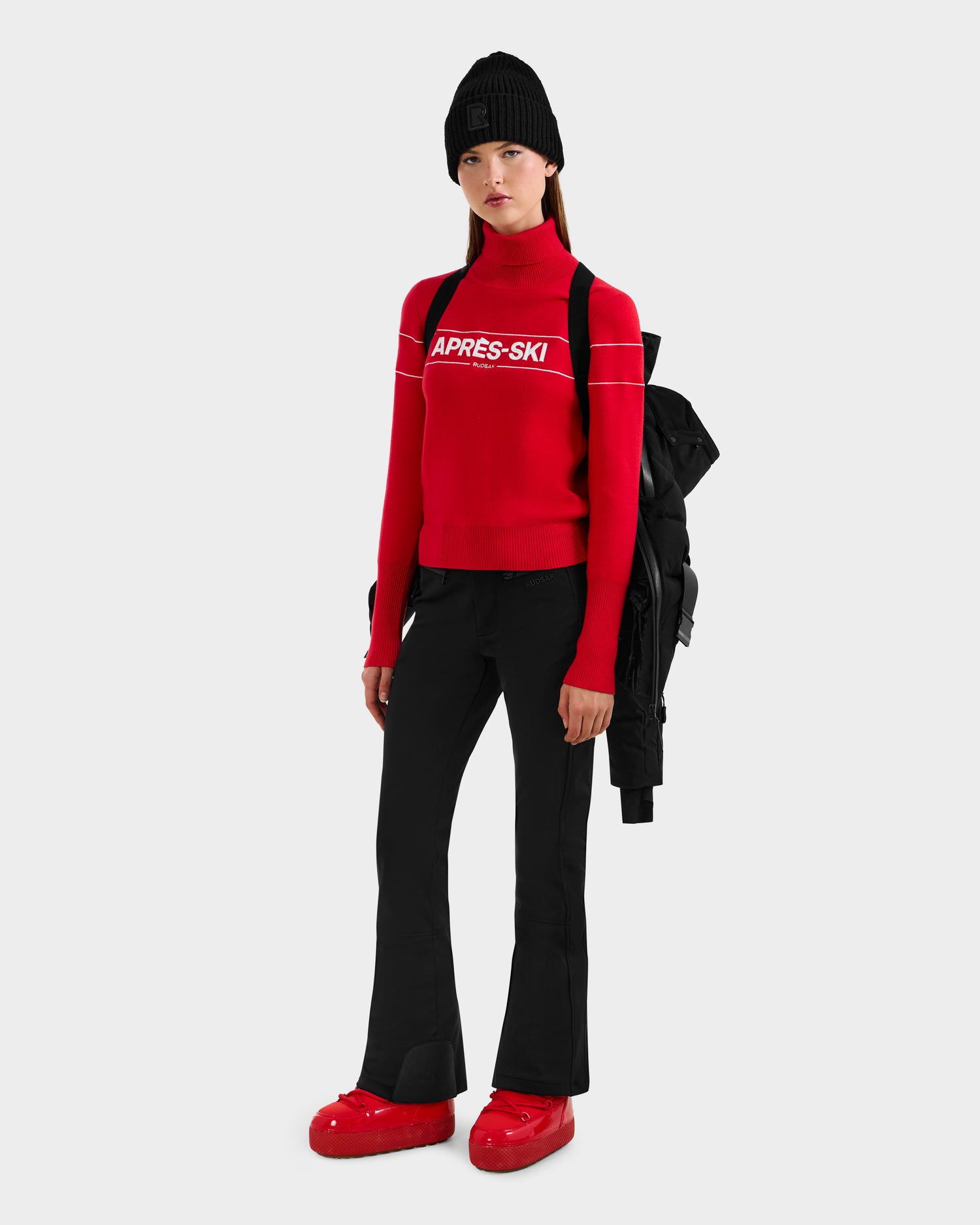 A woman in a black beanie, red APRÈS-SKI turtleneck, black pants, and red snow boots stands holding the Rudsak JOANNA - 8124788 Black women's down ski jacket with ski-pass pocket over her shoulder against a light gray background.