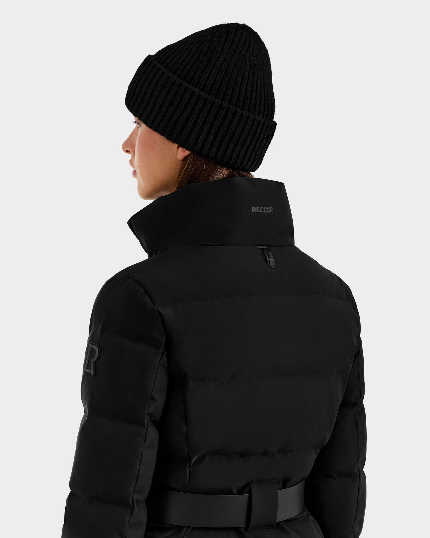 A person is seen from behind wearing the Rudsak JOANNA - 8124788 Black women's down ski jacket with a high collar and a black knit beanie, set against a plain light background.