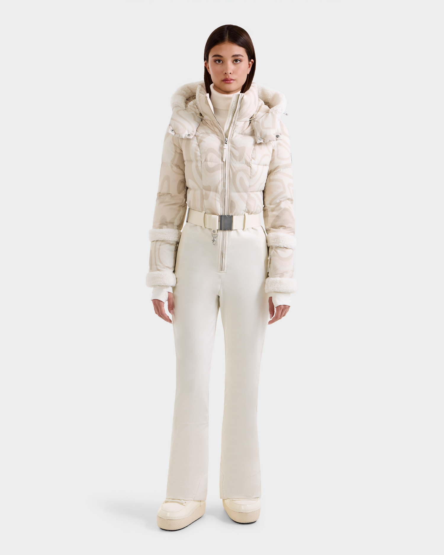 A woman faces forward in the Rudsak CARMEN P - 8125707 Vanilla Deja vu: a cream-patterned ski jacket with hood, matching high-waisted pants, and platform boots against a white background. Pre-order now; ships by November 30th.