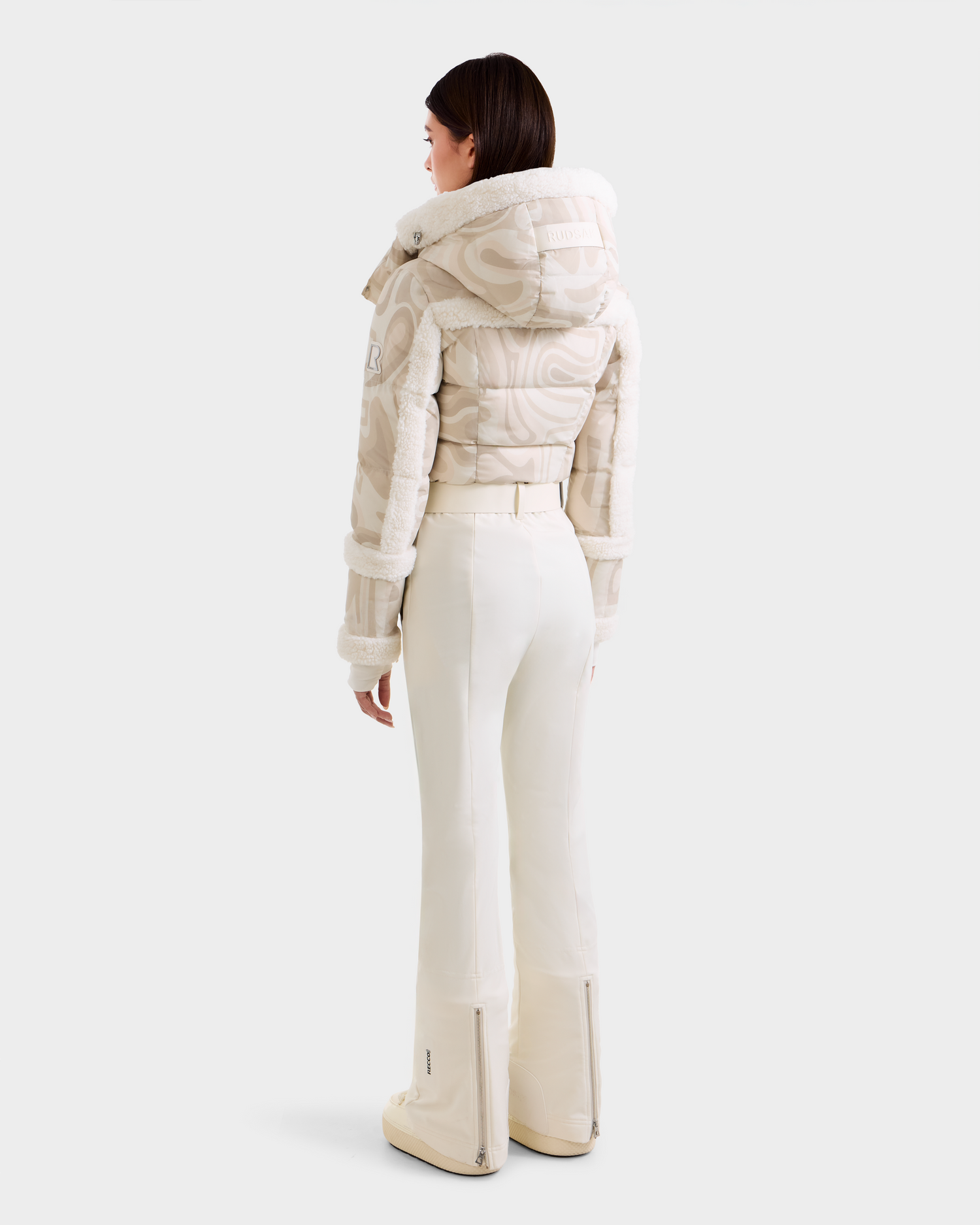 A woman faces away, wearing the Rudsak CARMEN P - 8125707 Vanilla Deja vu winter jacket and off-white flared pants with ankle zippers, against a light background. Pre-order now; ships by November 30th.