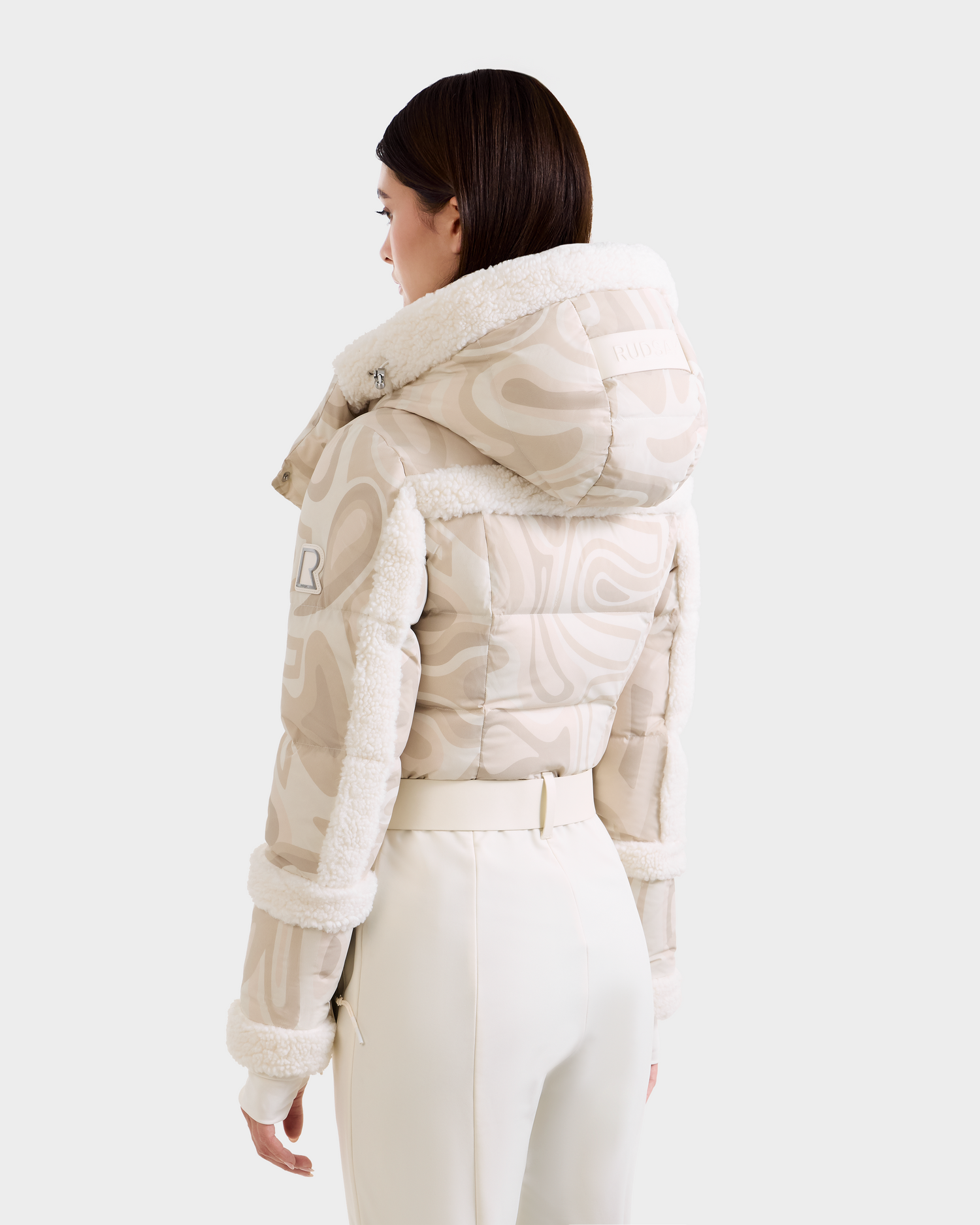 A woman faces away, wearing the Rudsak CARMEN P - 8125707 Vanilla Deja vu puffer jacket with shearling trim and hood, paired with high-waisted white pants on a light background. Available for pre-order; ships by November 30th.