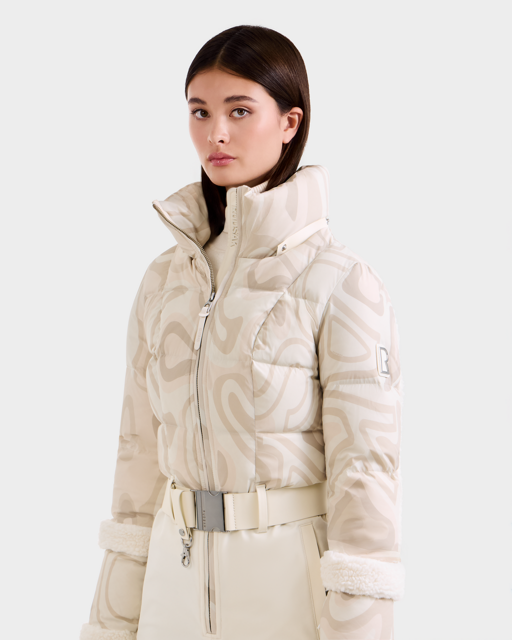 A woman with straight dark hair wears the Rudsak CARMEN P - 8125707 Vanilla Deja vu: a high-collared, patterned cream puffer jacket and matching belted pants with faux fur cuffs. Available for pre-order, ships by November 30th.