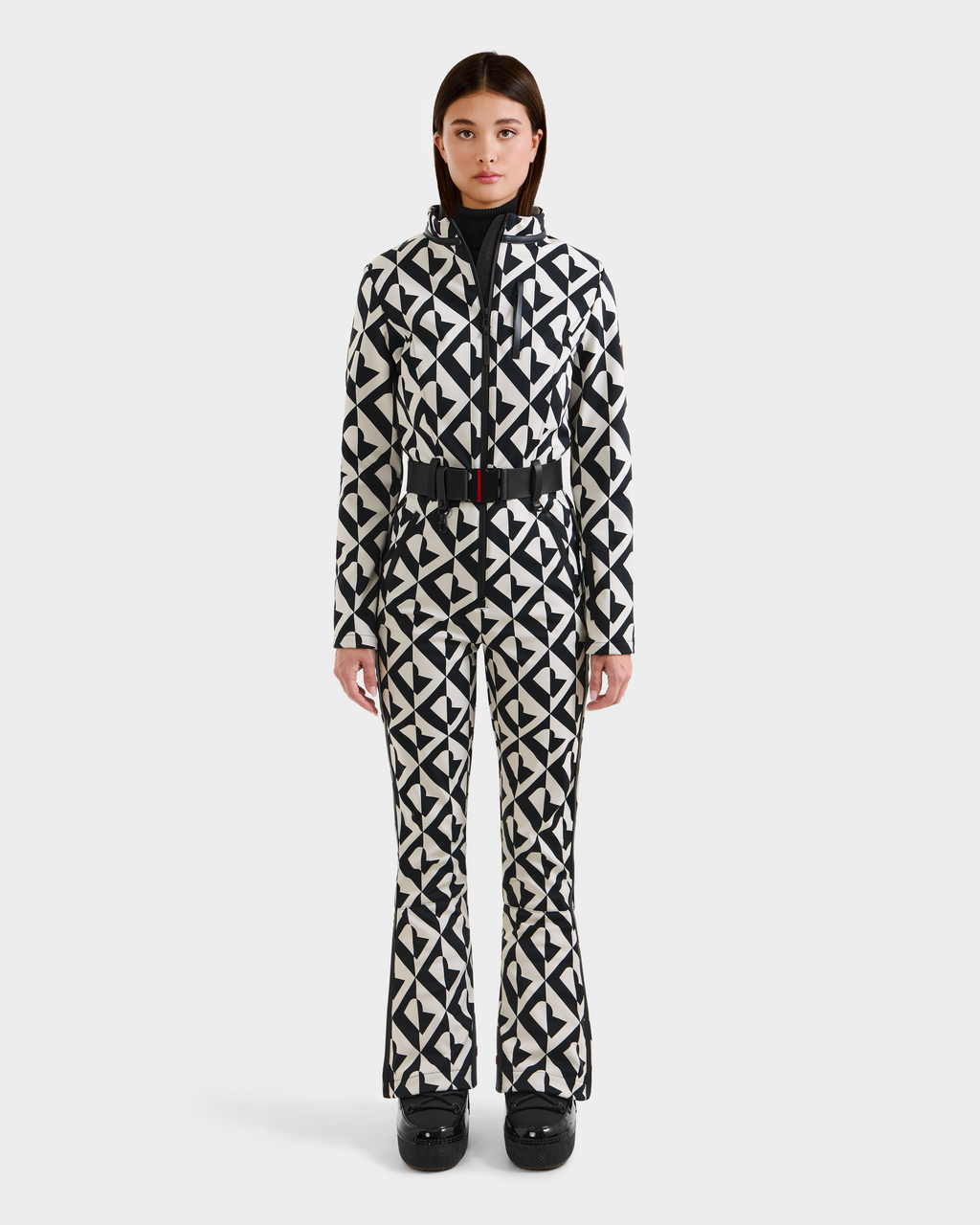 A woman faces forward in the Rudsak JOSIE - 8124817 Vanilla Monogram one-piece ski suit featuring a belted waist, flared pants, black and white monogram pattern, black ski boots, and RECCO rescue tech against a white background.