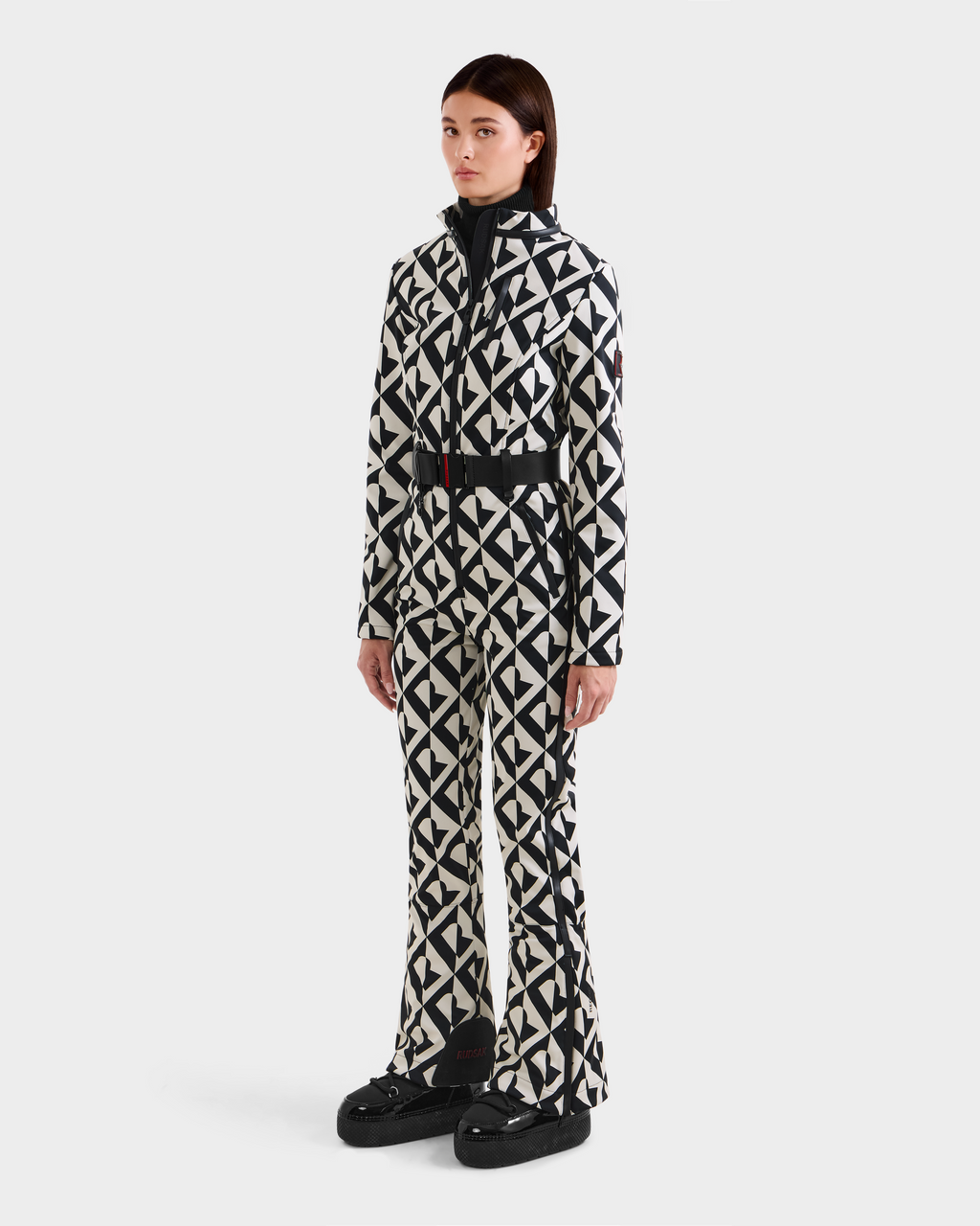 A woman poses against a white background wearing the Rudsak JOSIE - 8124817 Vanilla Monogram ski suit, featuring a matching black and white geometric pattern with flared legs and black platform shoes. She has straight, shoulder-length brown hair.