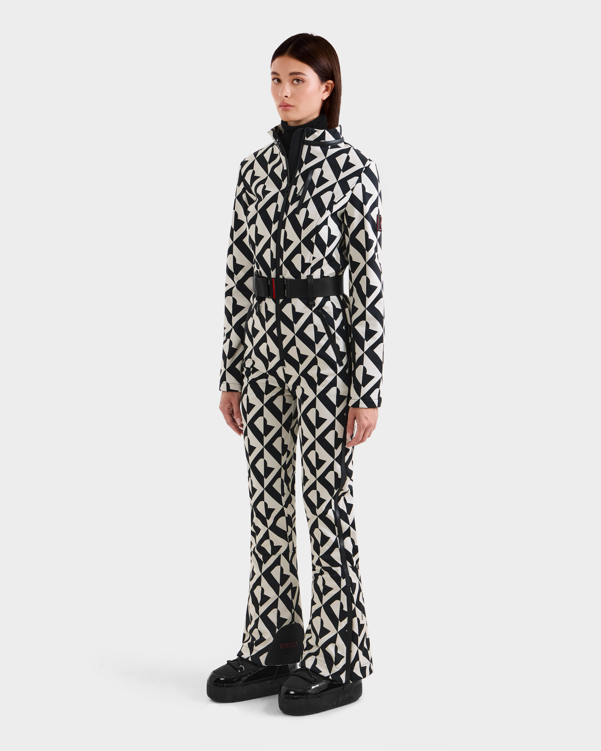 A woman poses against a white background wearing the Rudsak JOSIE - 8124817 Vanilla Monogram ski suit, featuring a matching black and white geometric pattern with flared legs and black platform shoes. She has straight, shoulder-length brown hair.
