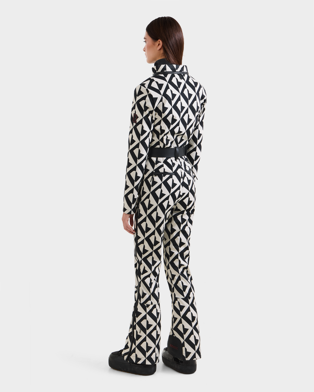 A person faces away, wearing the Rudsak JOSIE - 8124817 Vanilla Monogram women’s ski suit with flared legs and black boots, set against a plain white background.