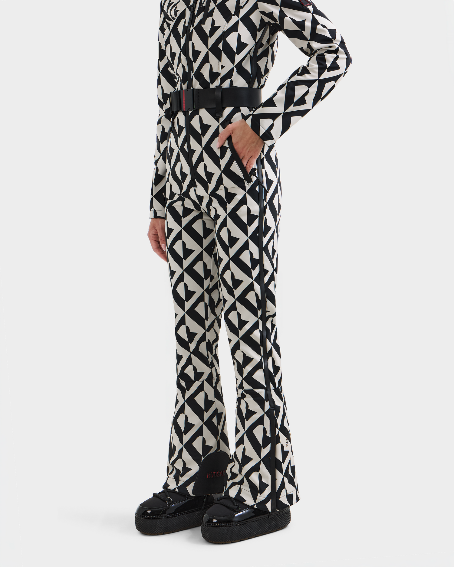 A person wearing the Rudsak JOSIE - 8124817 Vanilla Monogram one-piece ski suit, featuring a black and white geometric pattern and black belt, with black platform shoes. Shown from the shoulders down against a plain white background.