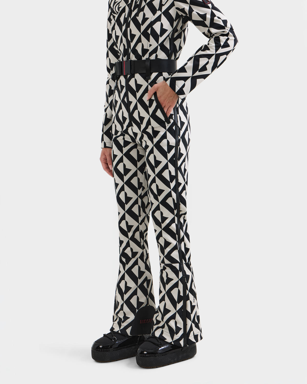 A person wearing the Rudsak JOSIE - 8124817 Vanilla Monogram one-piece ski suit, featuring a black and white geometric pattern and black belt, with black platform shoes. Shown from the shoulders down against a plain white background.