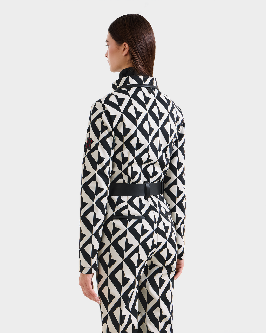 A woman with straight brown hair stands facing away, wearing the Rudsak JOSIE - 8124817 Vanilla Monogram women's ski suit with a black and white geometric pattern. The background is plain white.