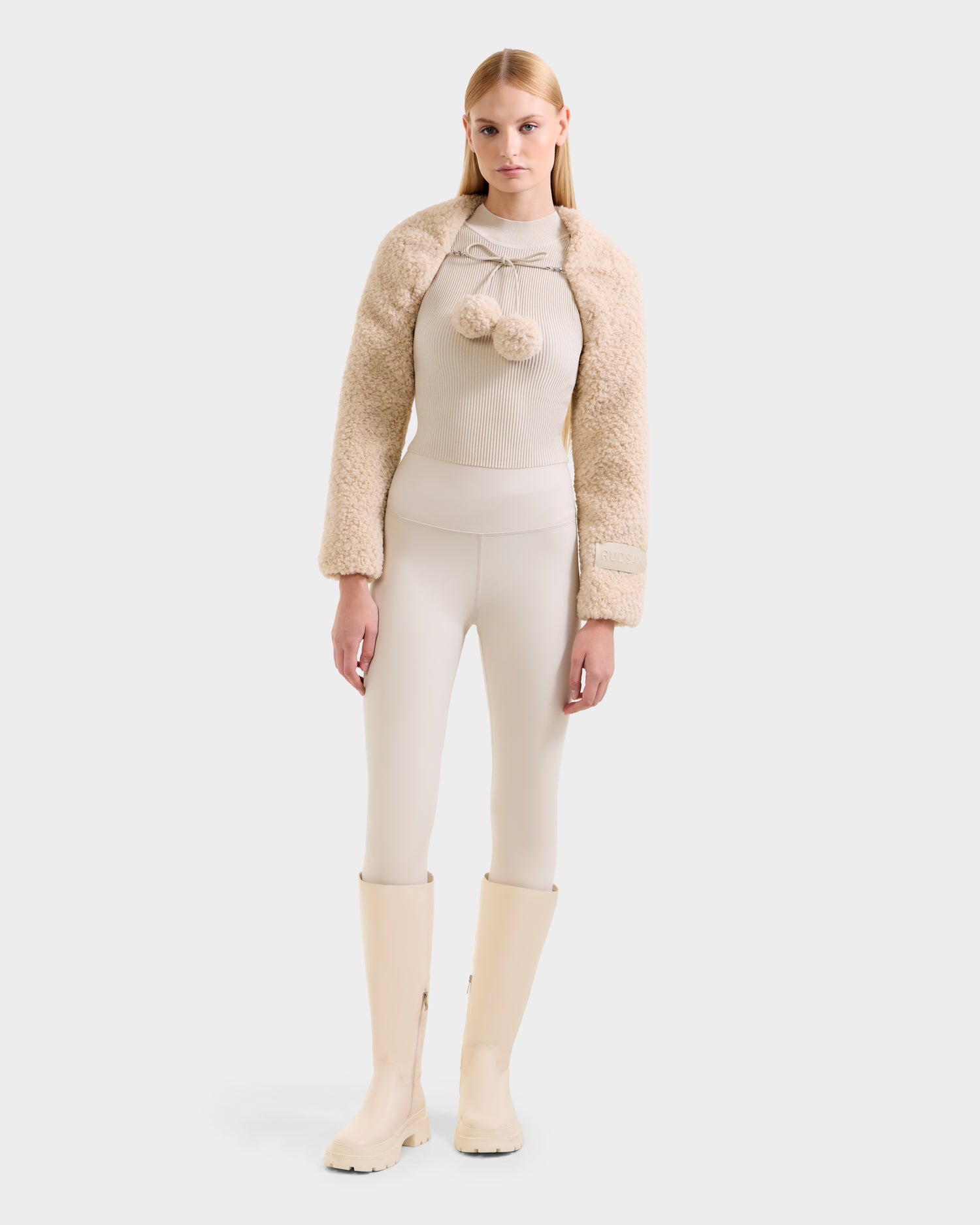 A woman faces forward in the Rudsak VERITY - 8525666 Vanilla fuzzy beige jacket against a plain white background, pairing it with cream high-waisted leggings, a fitted ribbed après-ski top with pom-poms, and knee-high cream boots. Her hair is straight and center-parted.