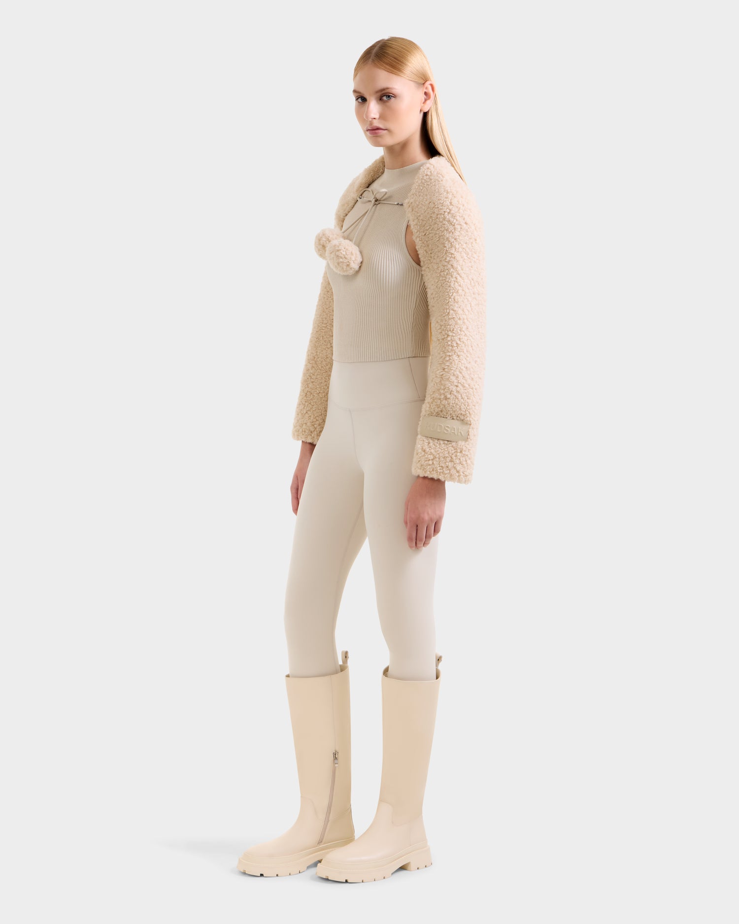 A woman with straight blonde hair models the Rudsak VERITY - 8525666 Vanilla jacket—an après-ski inspired beige textured piece—paired with cream separates and knee-high boots, standing against a plain light gray background.
