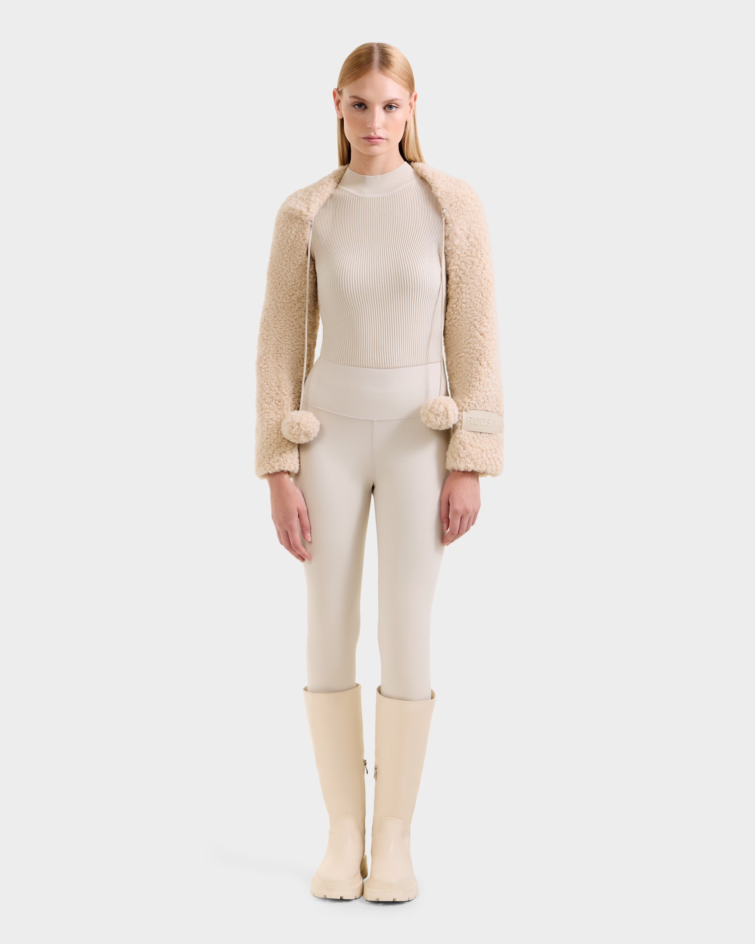 A woman faces forward in the Rudsak VERITY - 8525666 Vanilla faux shearling bolero over a cream ribbed turtleneck, fitted pants, and tall boots—an effortless cold-weather statement. The background is plain white.