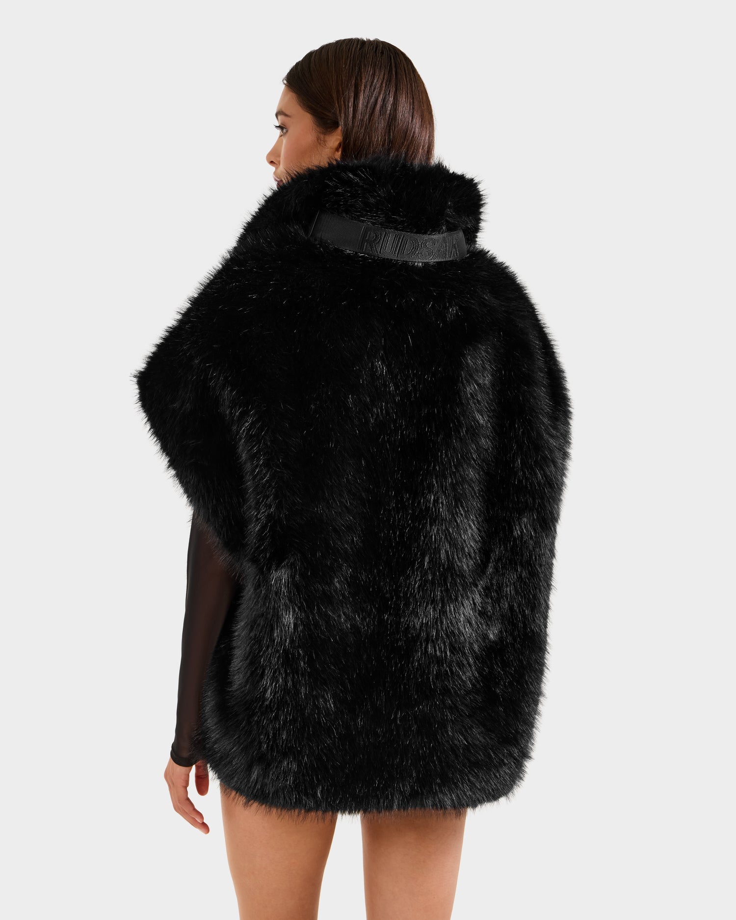 A woman with straight brown hair is seen from behind wearing the Rudsak DELFINA - 8525662 Black faux fur cape with an oversized silhouette over a black outfit, set against a plain light gray backdrop for chic winter style.