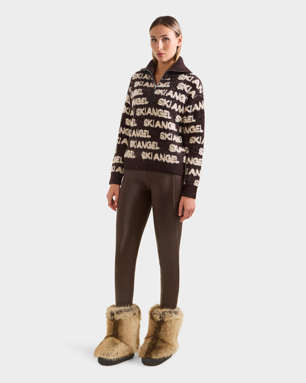 A woman wears the Rudsak LYDIA - 8525687 Espresso alpine pullover sweater with brown leggings and tan fur boots, her hair pulled back as she poses against a plain white background, perfect for après-ski.