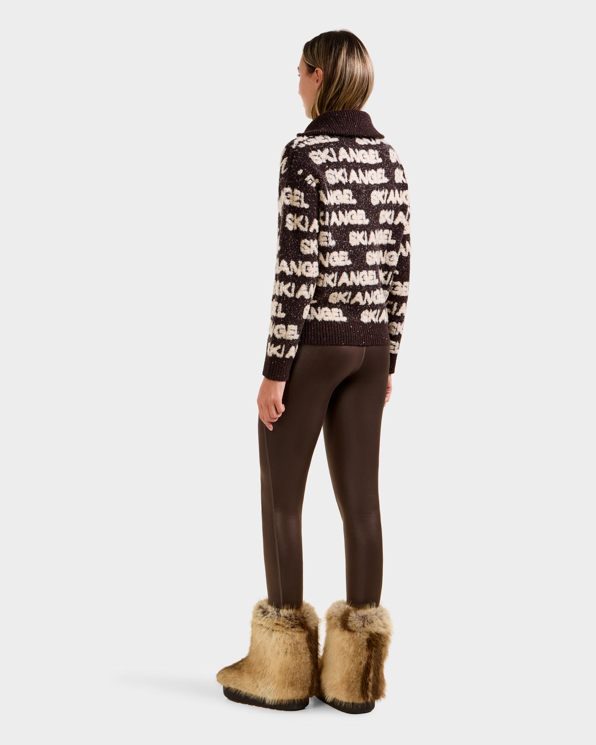 A person stands facing away, wearing the Rudsak LYDIA - 8525687 Espresso alpine pullover with SKI ANGEL text, paired with shiny brown leggings and tan furry boots. Their straight hair is loose against a plain white background.