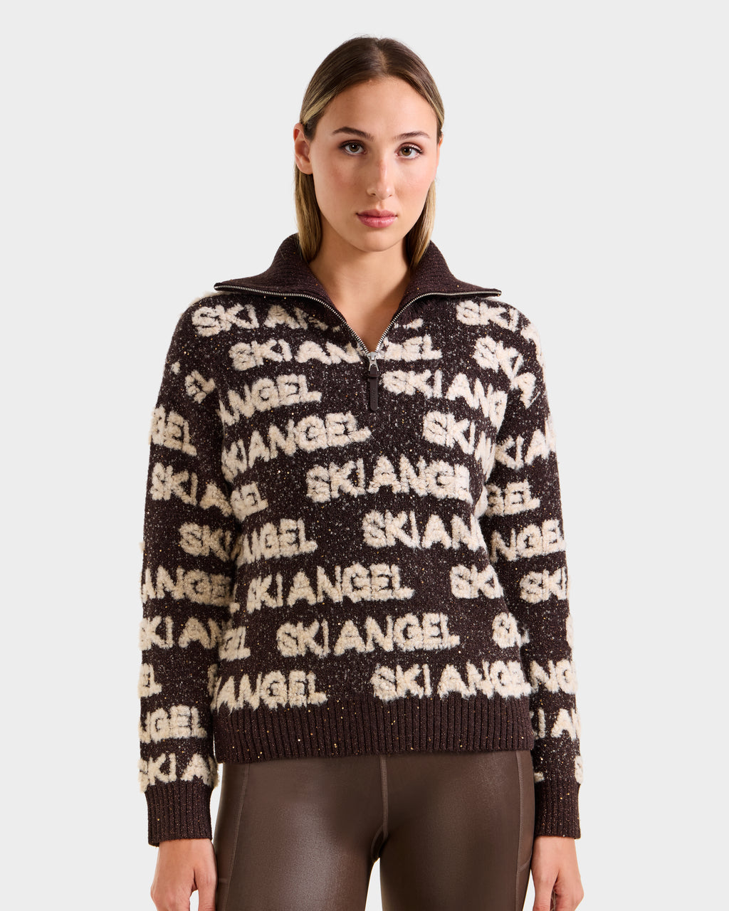 A woman with straight hair models the Rudsak LYDIA - 8525687 Espresso zip-up sweater featuring bold white SKI ANGEL print, paired with shiny brown pants—an ideal winter look—against a plain light background.