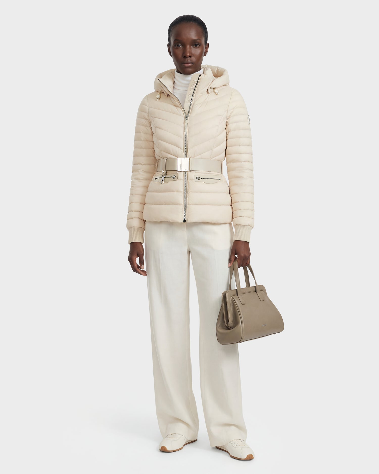 Against a plain background, a person wears the Rudsak SCARLETT - 8125004 Vanilla women's lightweight down jacket with a vegan leather waist belt, wide-leg white pants, white turtleneck and shoes, holding a taupe handbag in their left hand.