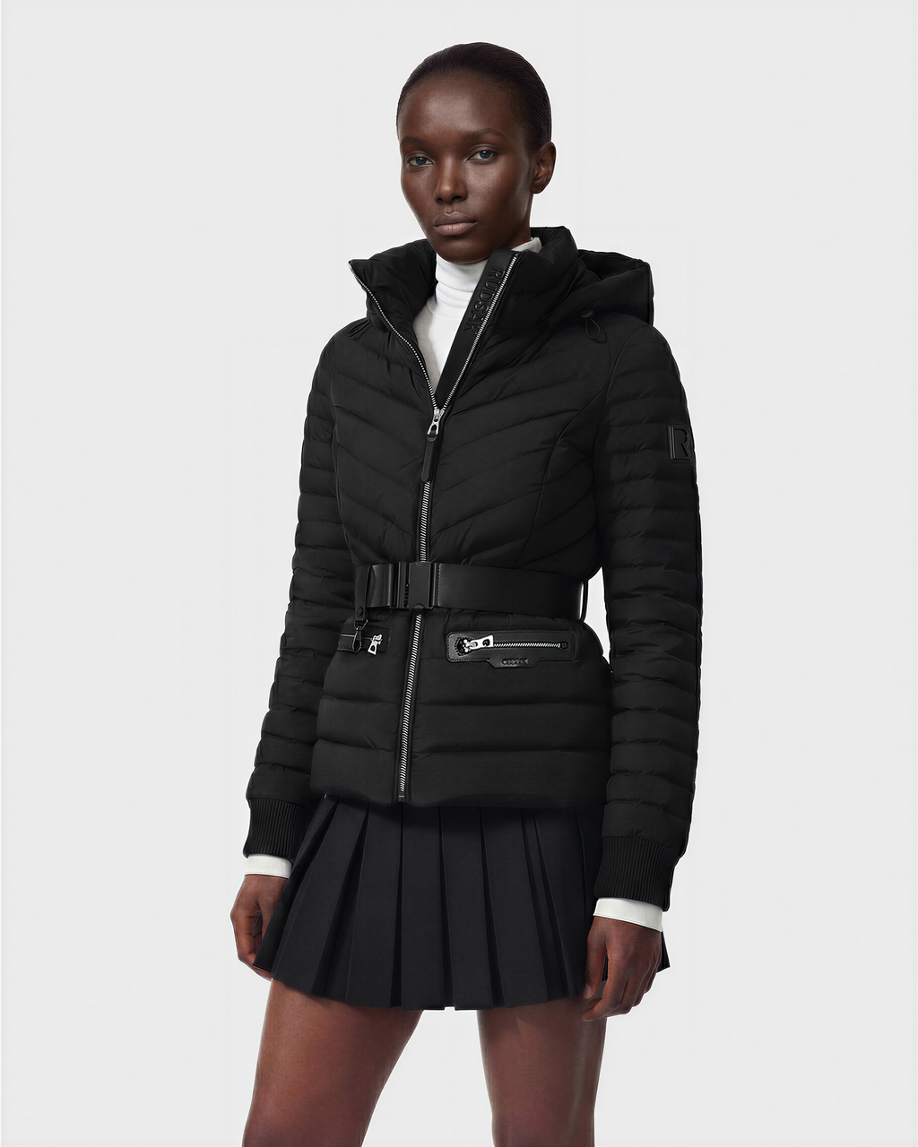 A person is wearing the Rudsak SCARLETT M - 8125565 Black lightweight down jacket, belted with zippered pockets, layered over a white turtleneck and black pleated skirt, standing against a plain light background.