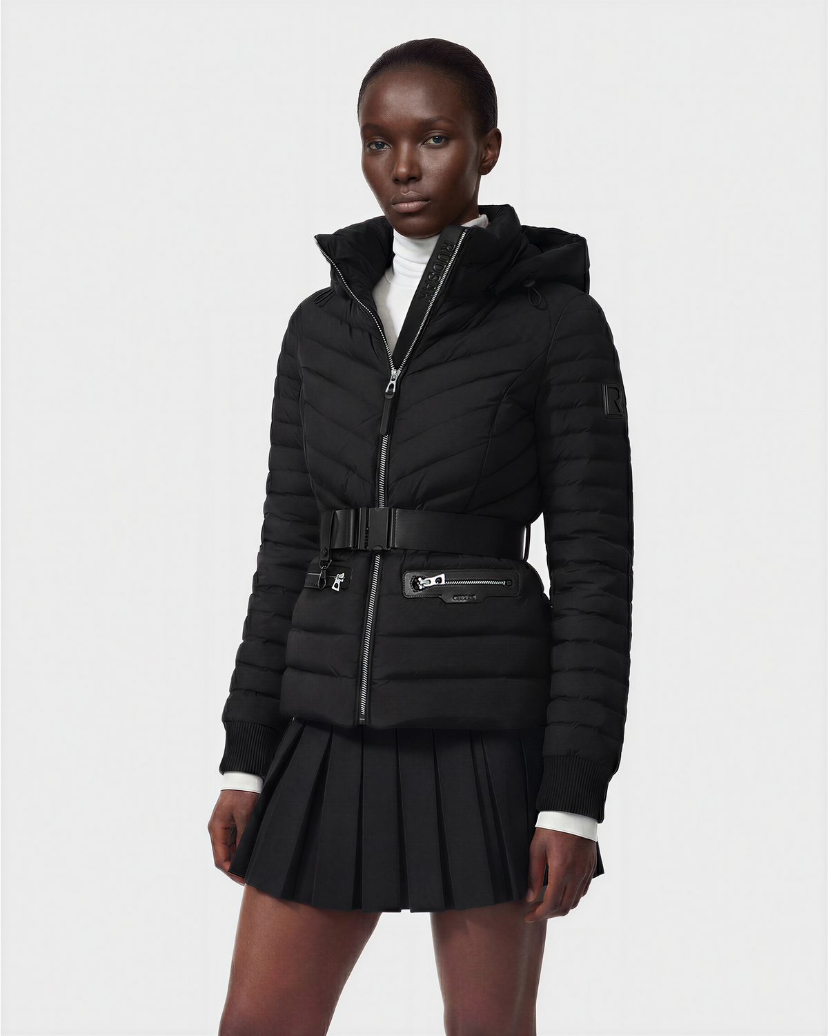 A person is wearing the Rudsak SCARLETT M - 8125565 Black lightweight down jacket, belted with zippered pockets, layered over a white turtleneck and black pleated skirt, standing against a plain light background.