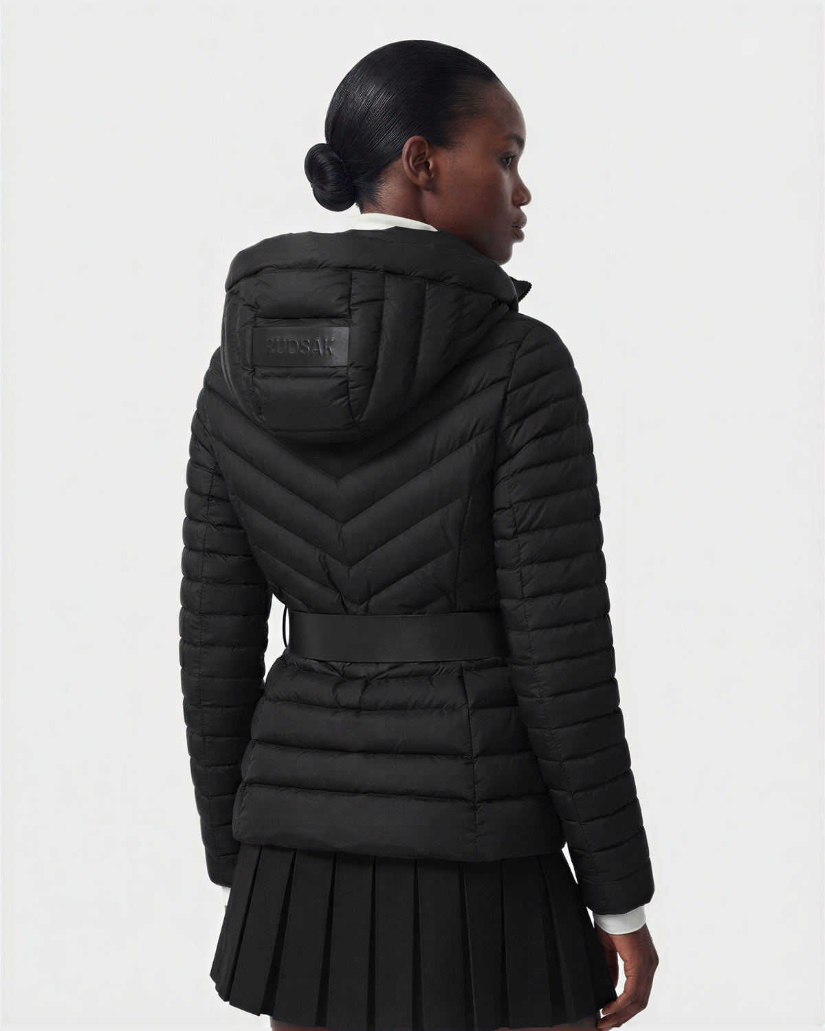 A woman, hair in a low bun, faces away wearing the Rudsak SCARLETT M - 8125565 Black lightweight down jacket with hood and a pleated black skirt. The background is plain and light-colored.