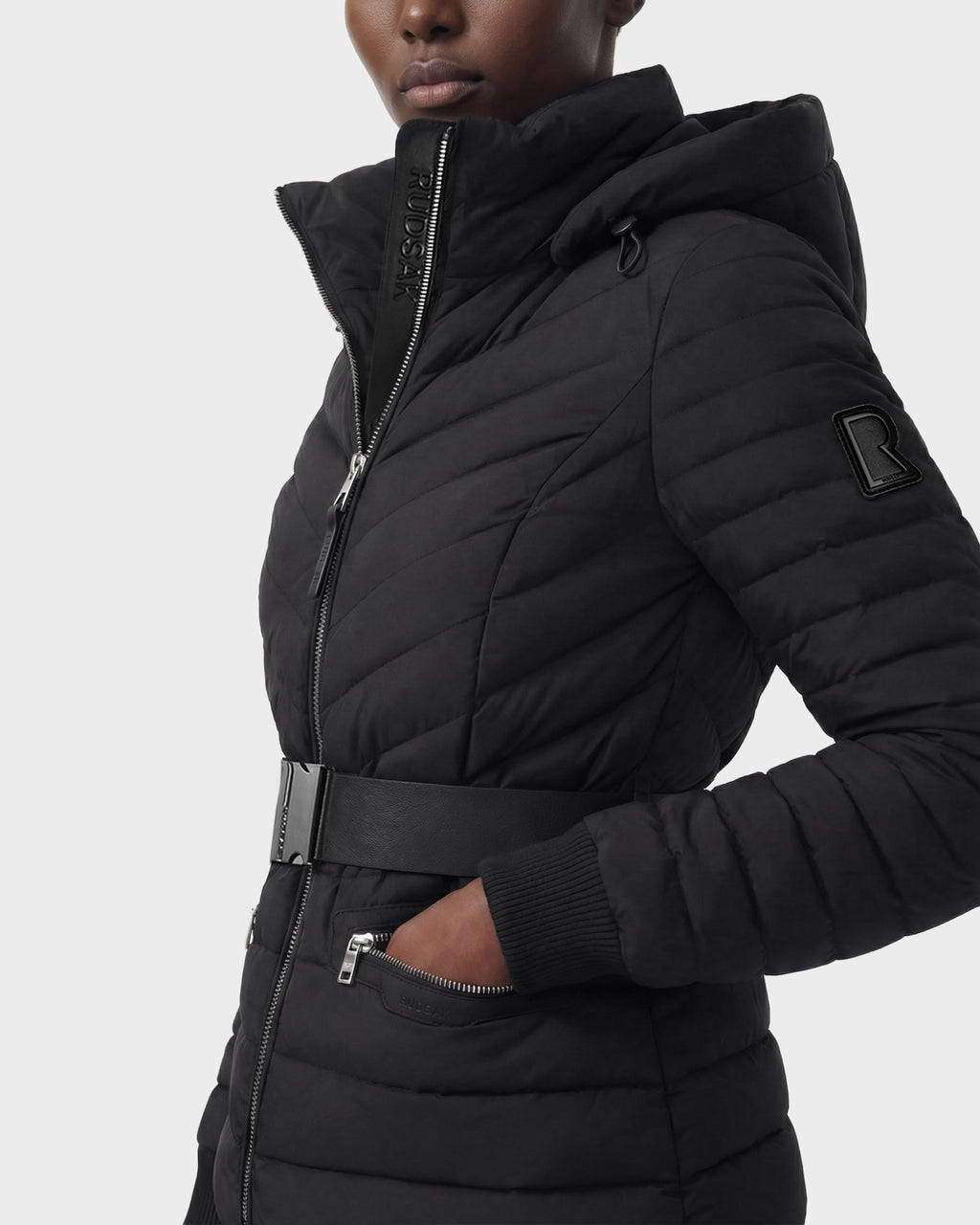A woman wears the Rudsak SCARLETT M - 8125565 Black quilted lightweight down jacket with a high collar, zipper front, belted waist, side pocket, and R patch on the sleeve. Made with RDS-certified down. Plain white background.
