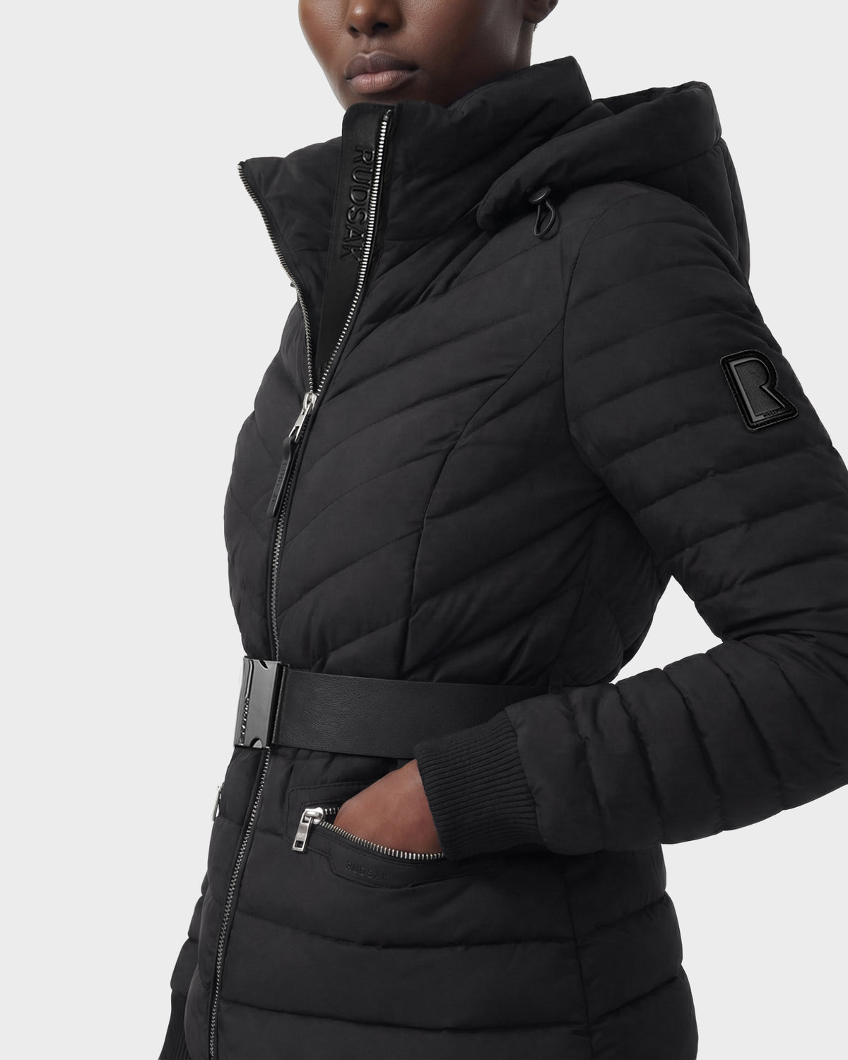 A woman wears the Rudsak SCARLETT M - 8125565 Black quilted lightweight down jacket with a high collar, zipper front, belted waist, side pocket, and R patch on the sleeve. Made with RDS-certified down. Plain white background.