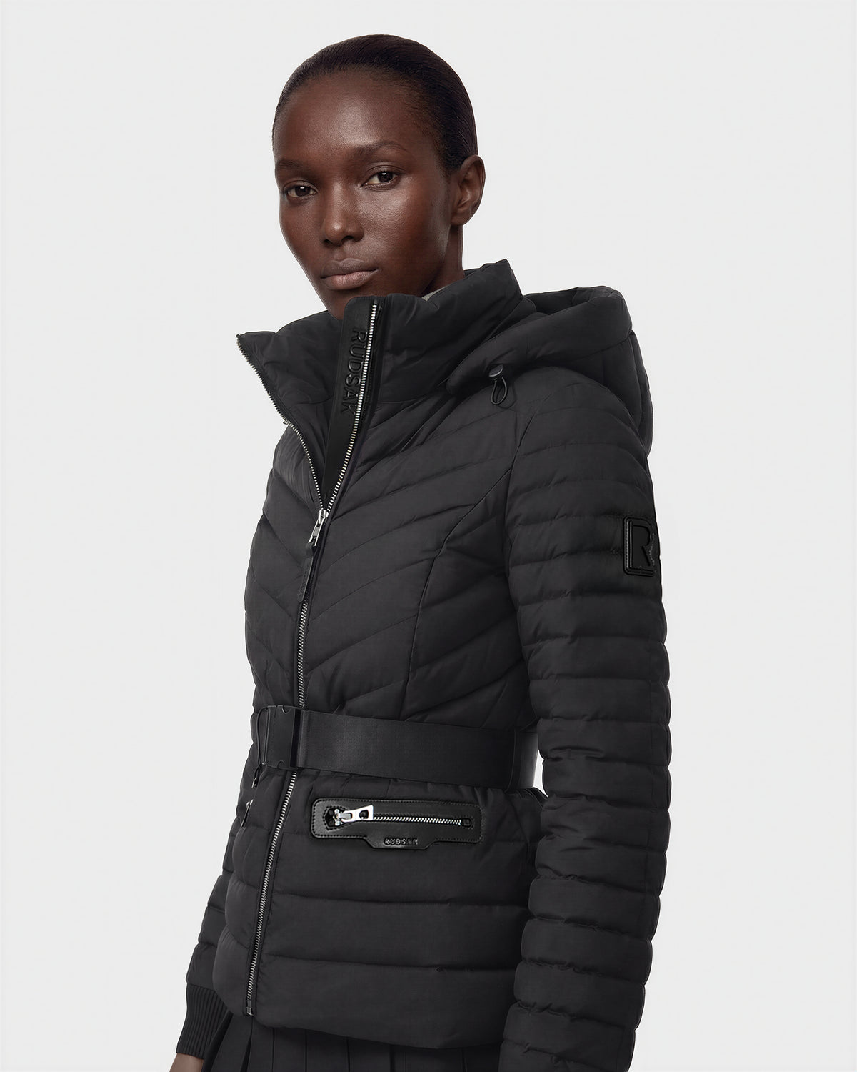 A person models the Rudsak SCARLETT M - 8125565 Black, a fitted women's lightweight down jacket with a vegan leather waist belt, high collar, and zippered pockets, standing against a plain white background and looking slightly to the side.