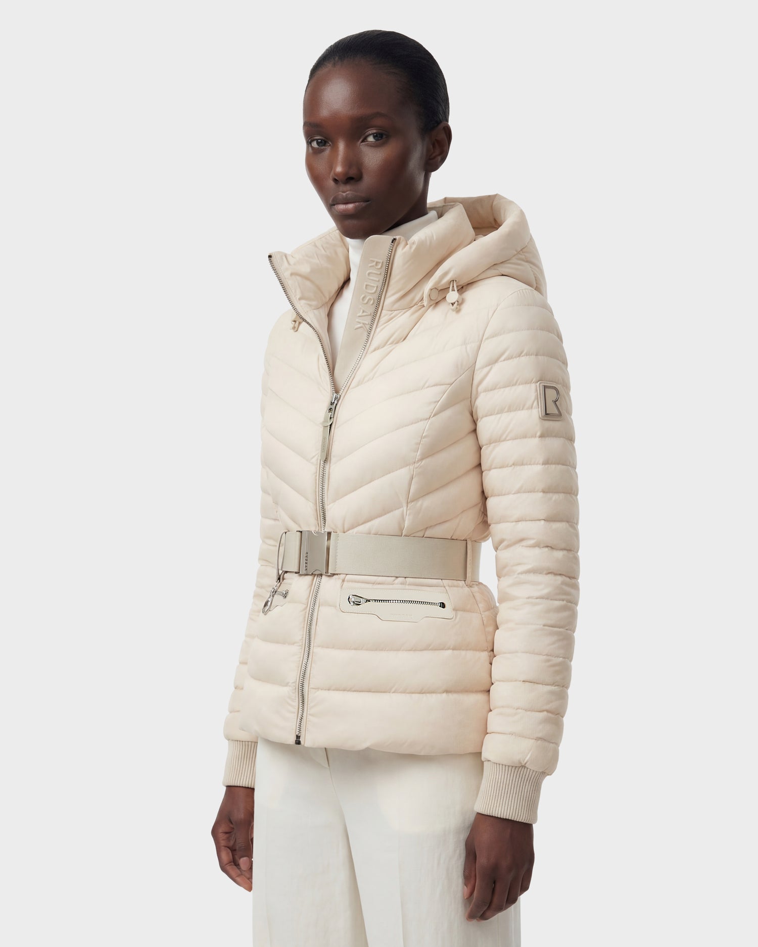 A woman wears the Rudsak SCARLETT - 8125004 Vanilla, a light beige quilted women’s lightweight down jacket with a vegan leather belt and zippered pockets, paired with white pants against a plain light background.