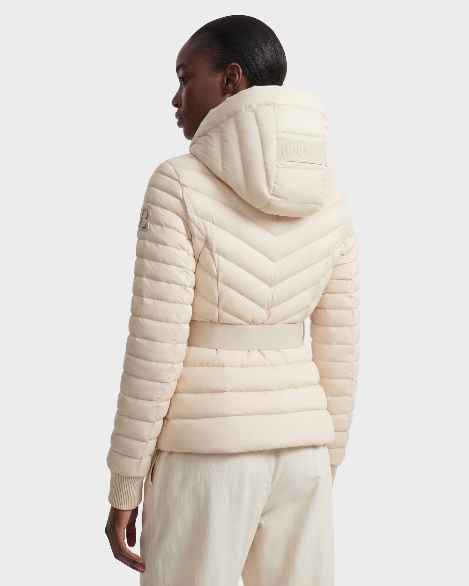 A person wears the Rudsak SCARLETT - 8125004 Vanilla women's lightweight down jacket in light beige with chevron stitching and embossed hood, crafted from RDS-certified down, paired with light-colored pants, shown from the back.