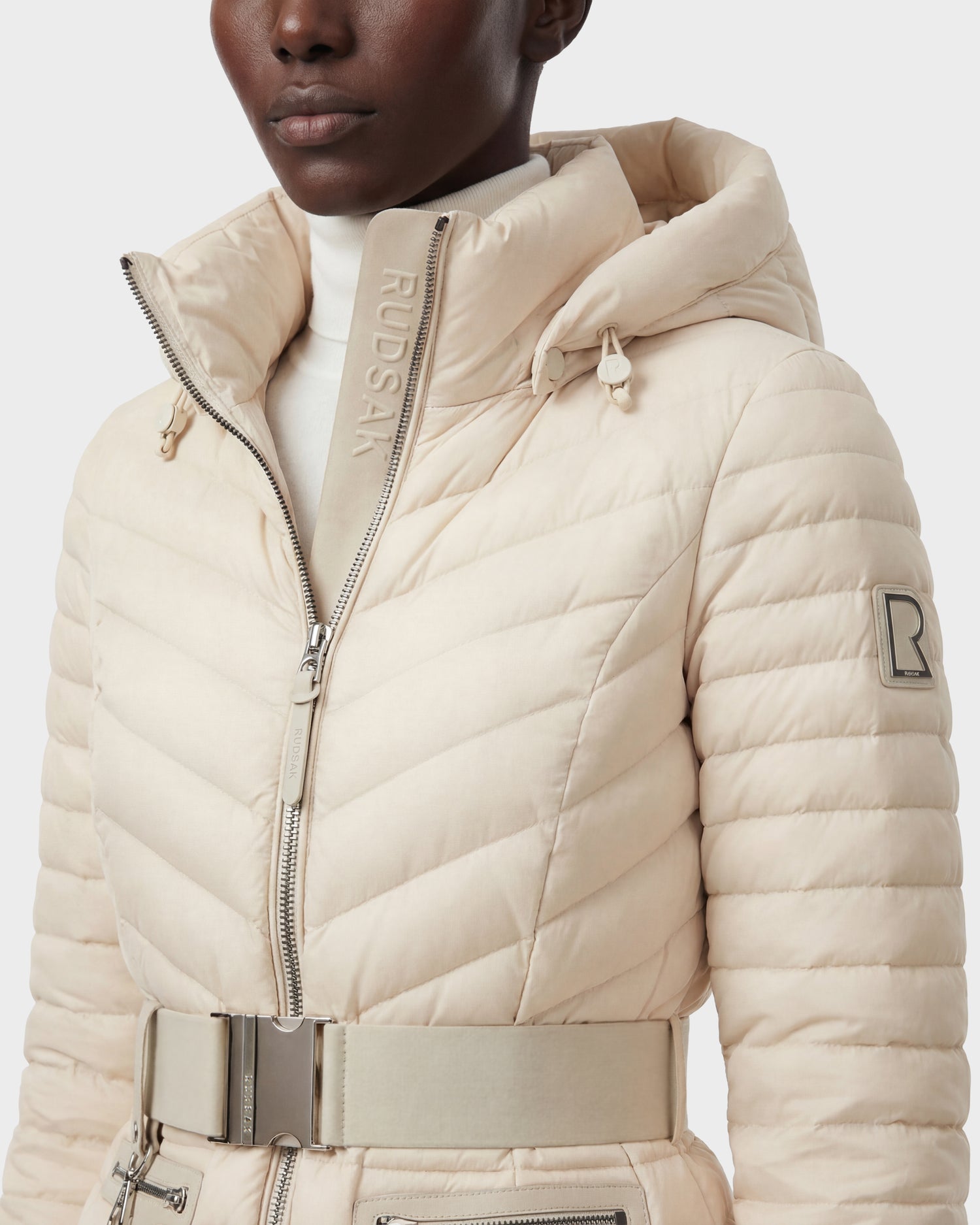 A person wears Rudsak's SCARLETT - 8125004 Vanilla, a cream quilted puffer jacket with a high collar, hood, vegan leather belt, visible zippers, and RUDSAK branding on the collar and sleeve.